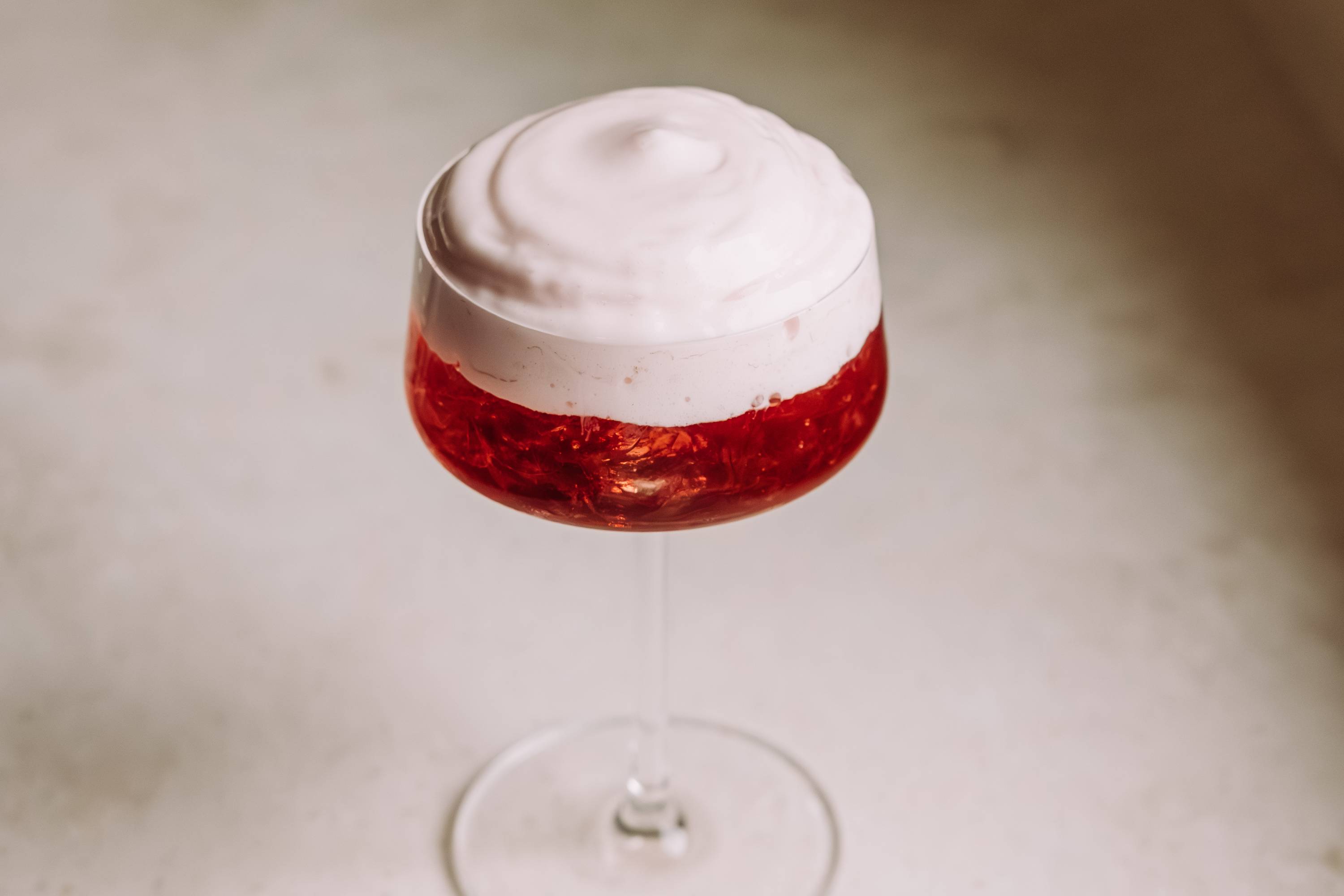 Transparentes Cocktailglas mit rotem Getränk, Eiswürfeln und weißer Schaumkrone