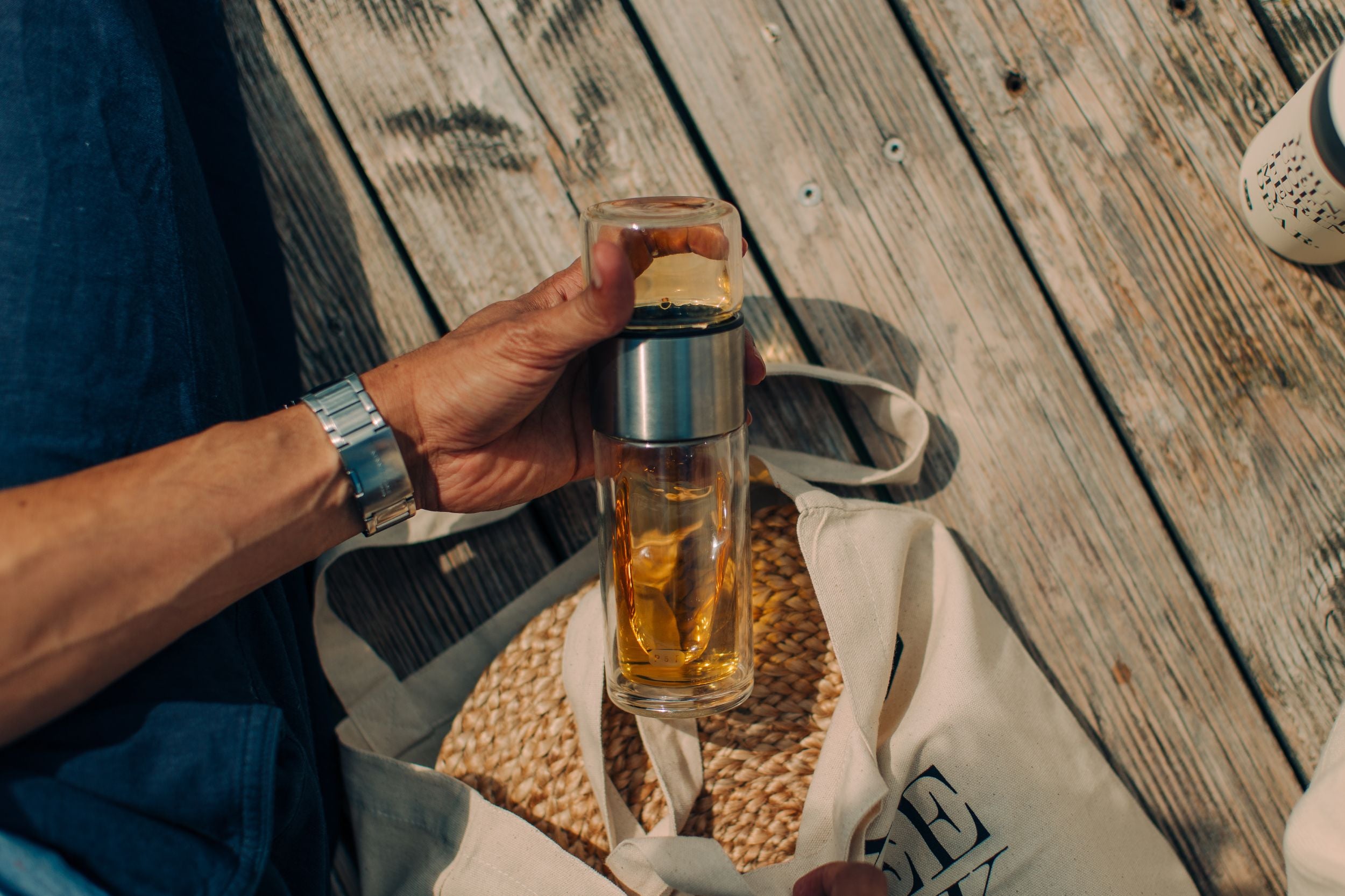 Person hält Glasflasche mit Edelstahl-Verzierung neben beigem Tote Bag
