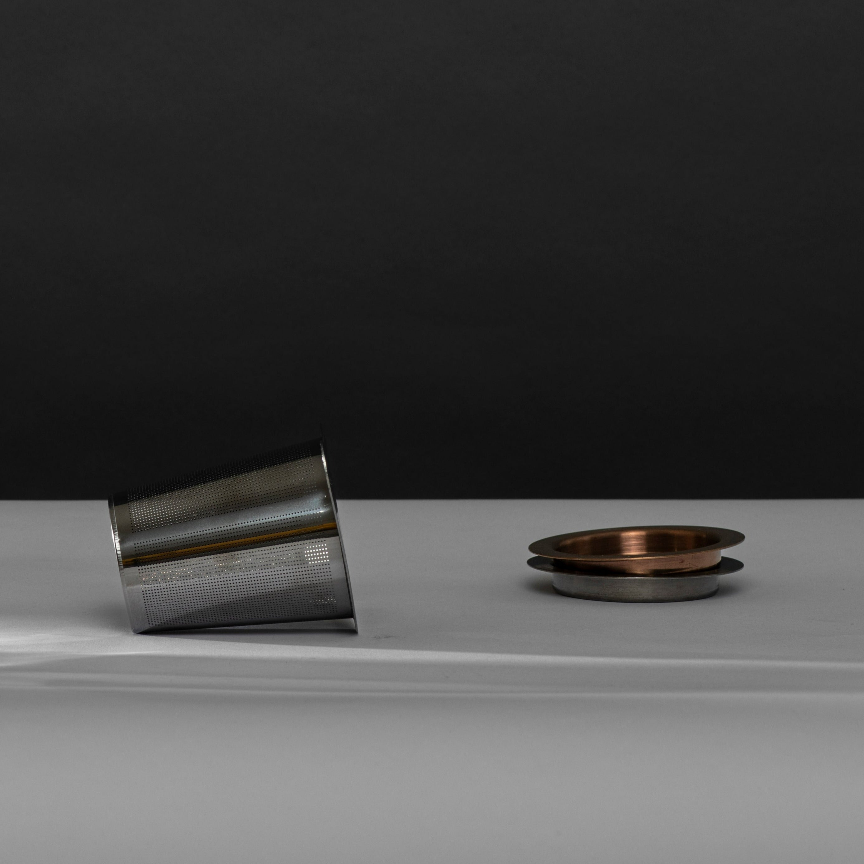 Silberner Teesiebzylinder mit Perforationen, gestapelten Metalldeckeln in Kupfer und Silber auf weißem Untergrund.