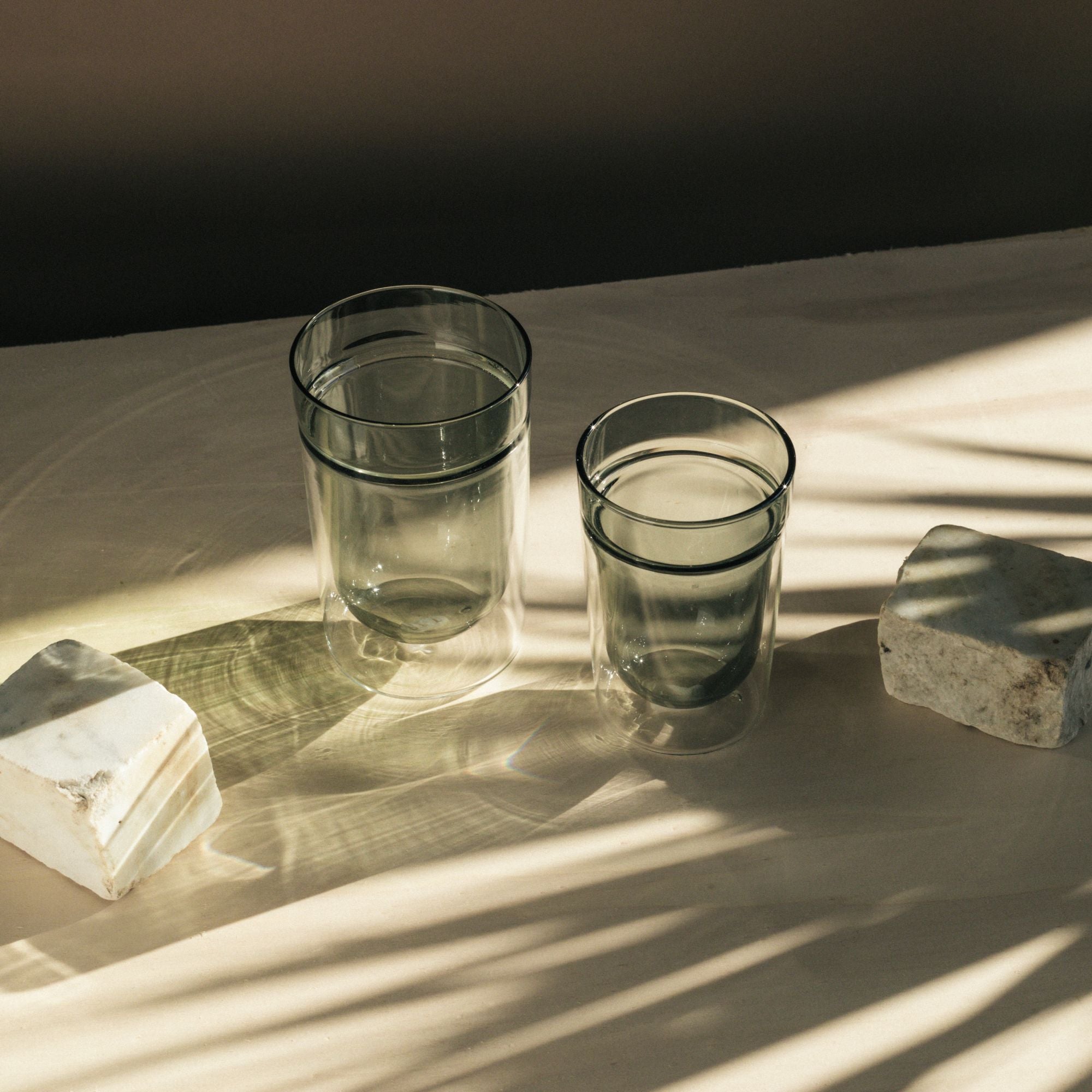 Zwei transparente Gläser mit Wasser auf beigem Untergrund, flankiert von weißen Steinen im sanften Licht.