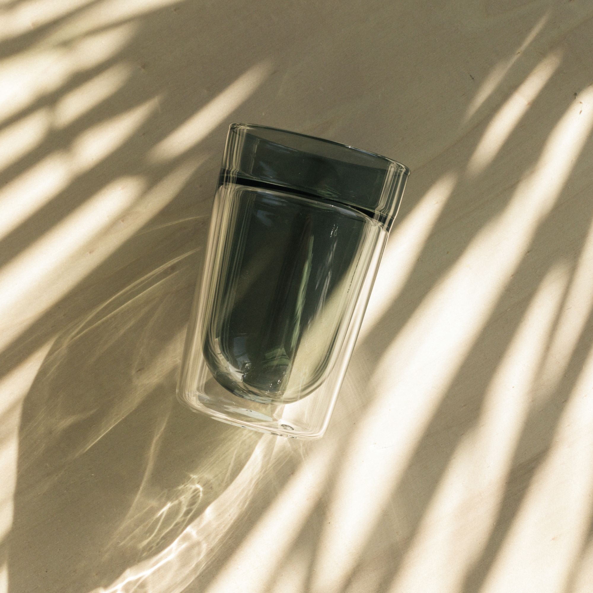 Doppeltwandiges Glas mit getöntem Inneren auf hellem Untergrund mit Palmblatt-Schattenmuster.