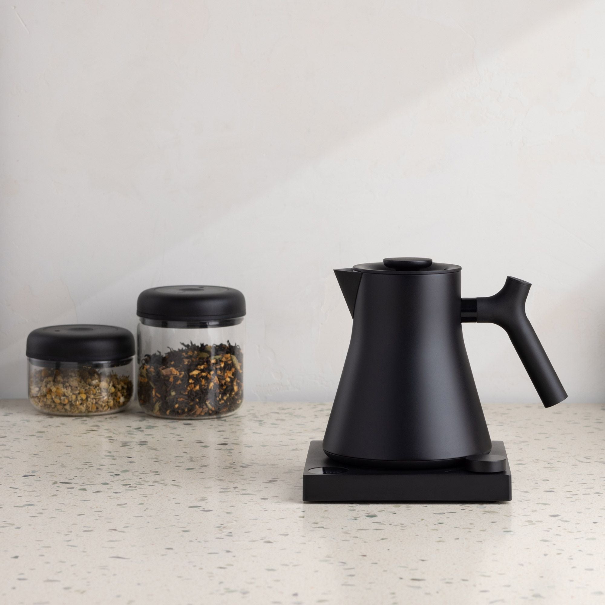 Moderner mattierter schwarzer Wasserkocher und zwei Gläser mit getrockneten Teeblättern auf Terrazzo-Arbeitsplatte.