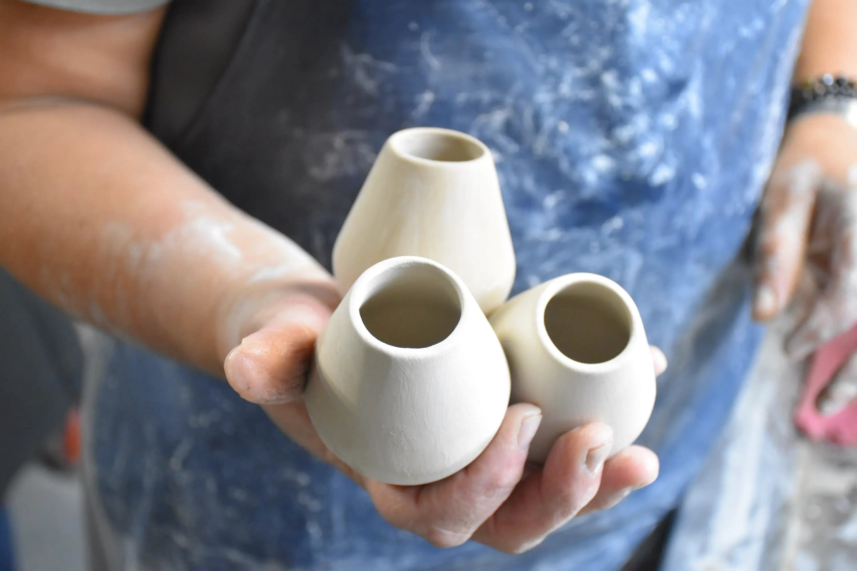 Hand hält drei unfertige, off-weiße Keramikbecher, mit Tonstaub bedeckt, symbolisiert Handwerkskunst.