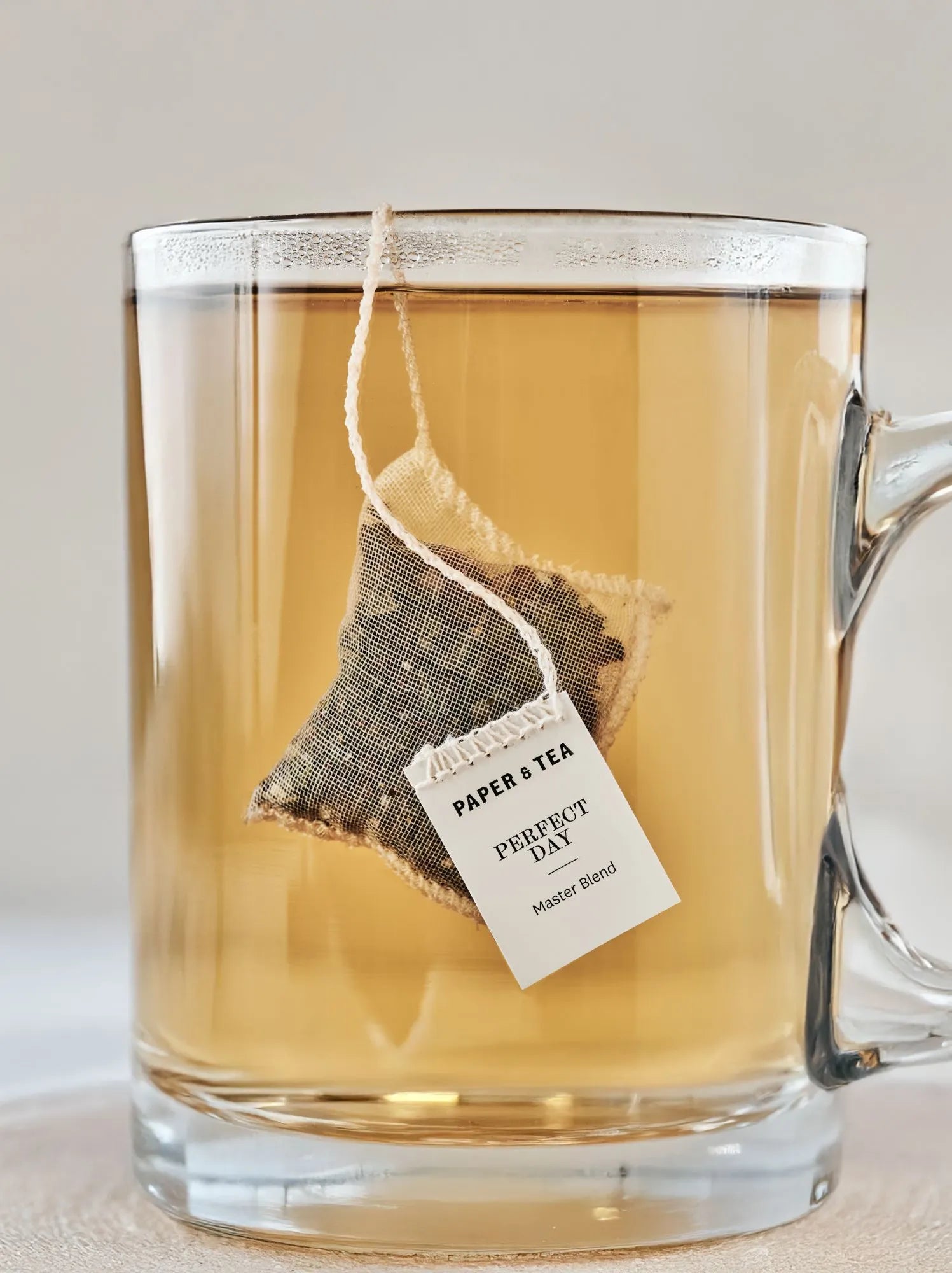 Klarer Glasbecher mit bernsteinfarbenem Tee und Mesh-Teebeutel von Paper & Tea.