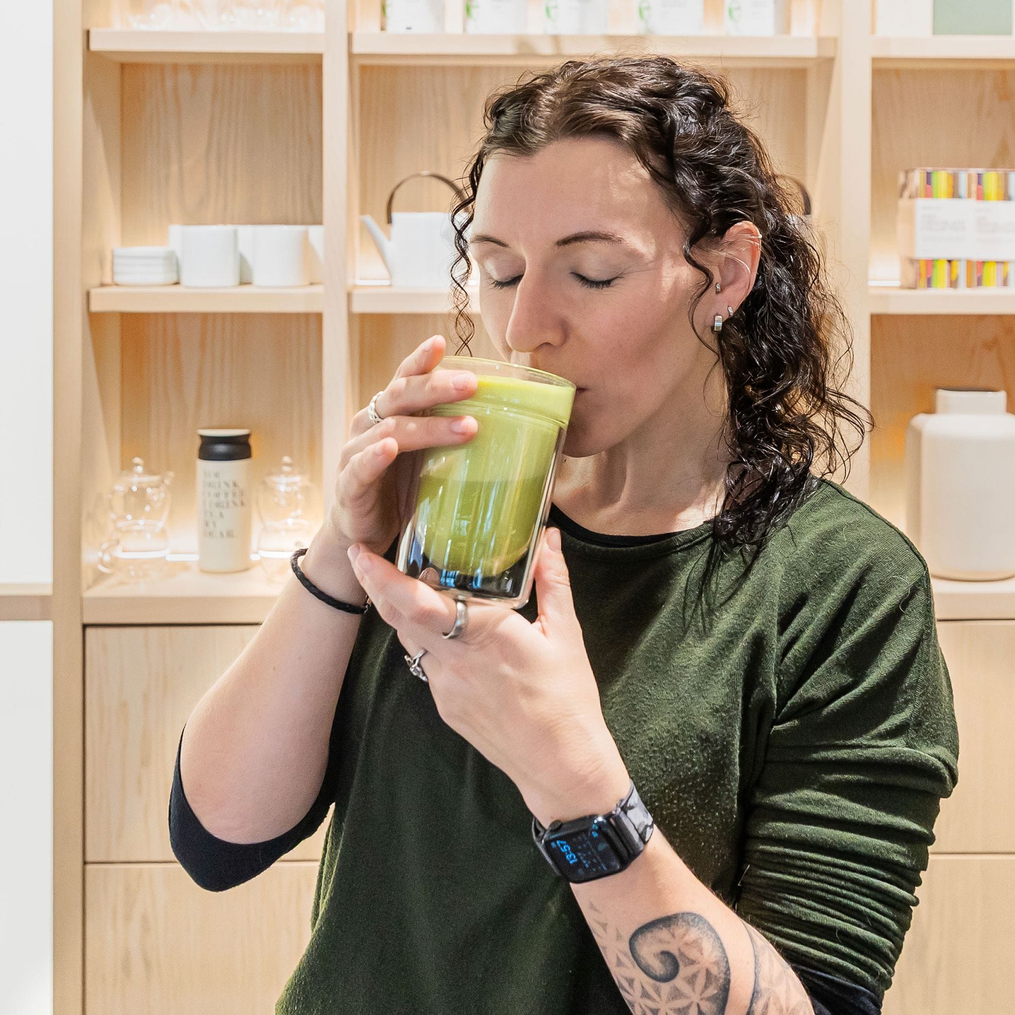 Person mit lockigem dunklem Haar trinkt grünen Smoothie aus Glas vor Holzregal mit Geschirr.
