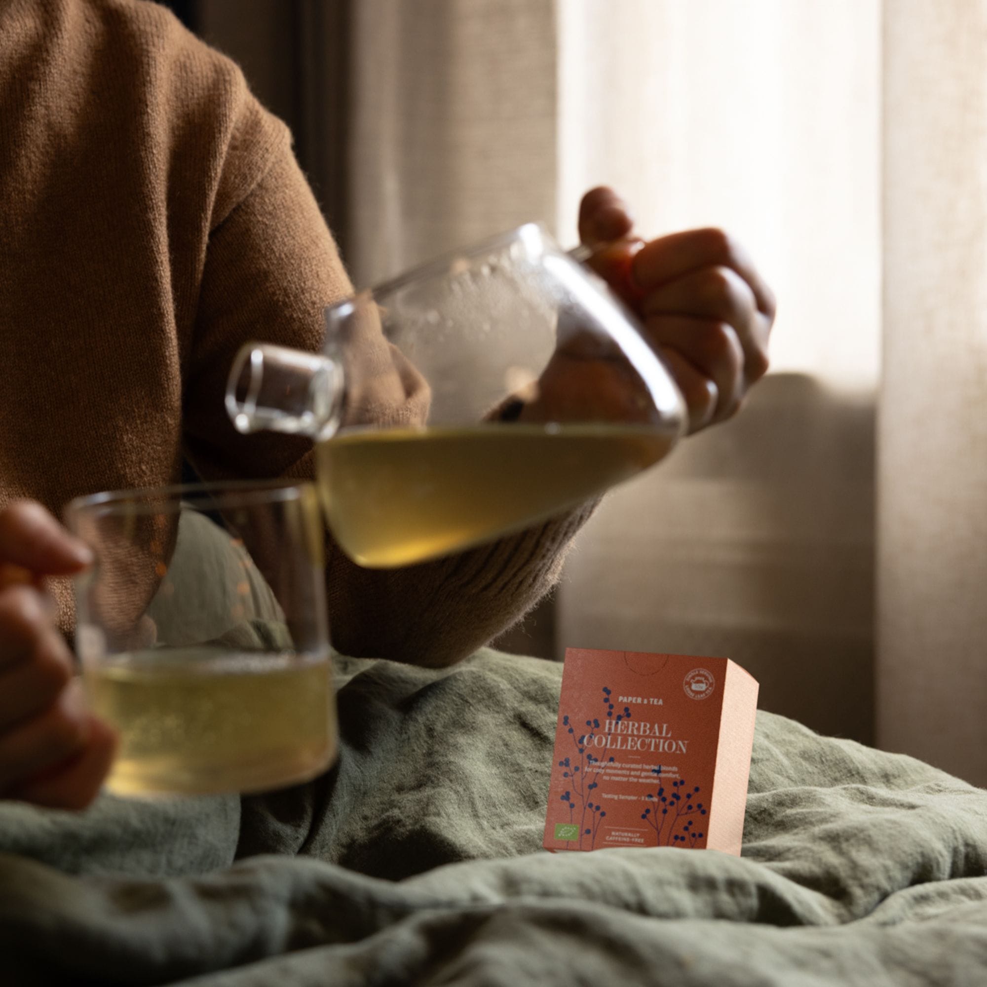 Person in braunem Pullover gießt Kräutertee aus Glas-Teekessel in Tasse neben PAPER & TEA Box.