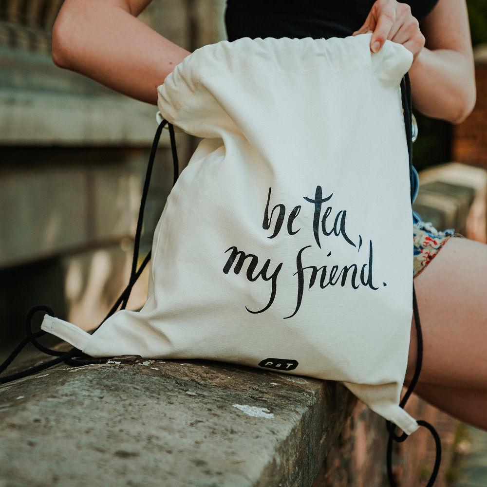 Weiße Stofftasche mit schwarzer Kordel und handgeschriebener Aufschrift "be tea, my friend.", gehalten von Person in schwarzem Oberteil und floralen Shorts auf einer steinernen Bank.