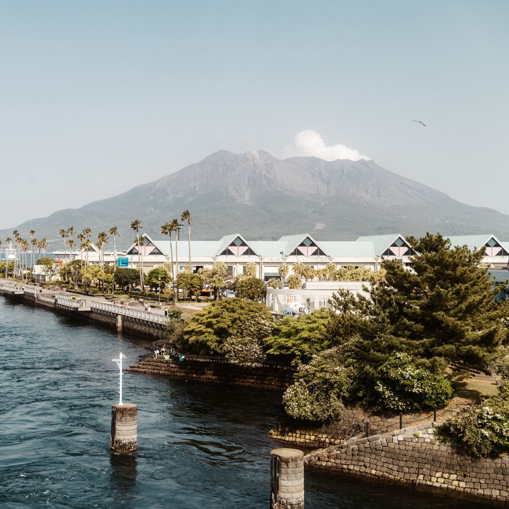 Szenische Uferpromenade, Vulkan im Hintergrund, pastellfarbenes Gebäude, grüner Park, blaues Wasser mit Boje.