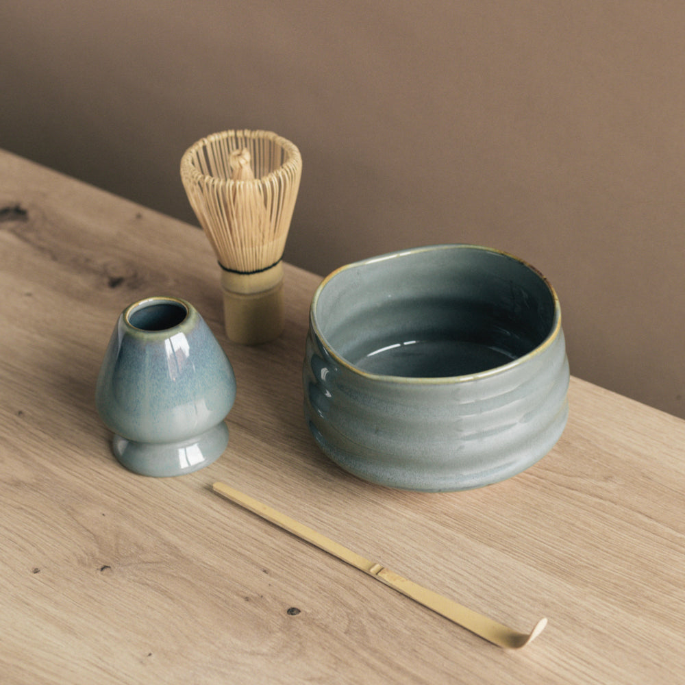 Minimalistisches, blau-graues Keramik-Teeservice mit Bambusbesen und Löffel auf hellem Holz vor brauner Wand.