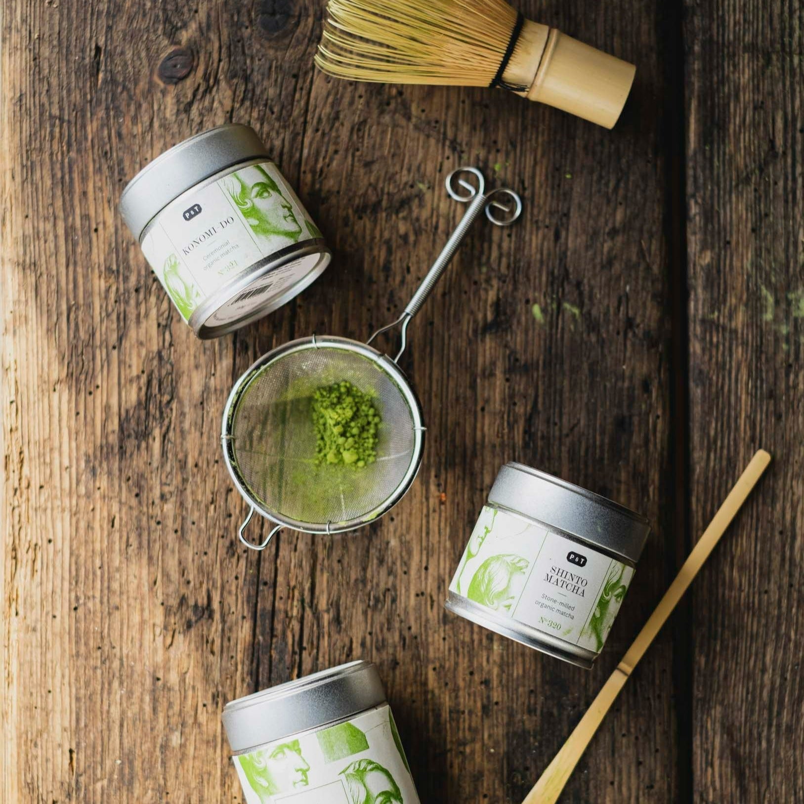 Zwei Dosen mit Bio-Matcha und steingemahlenem Matcha von P&T auf Holz, mit Sieb, Bambusbesen und Löffel.
