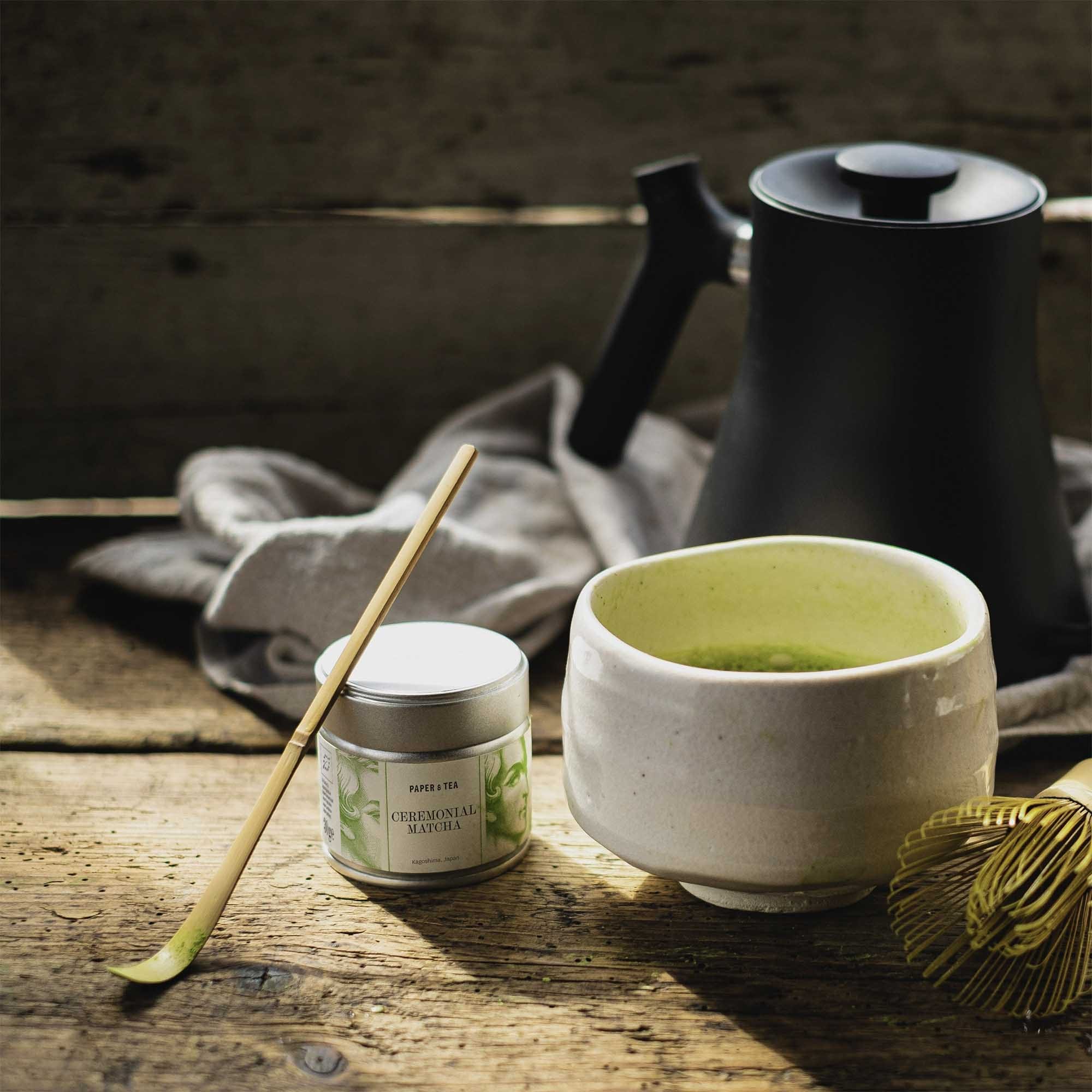 Weißer Keramikbecher mit leuchtend grünem Matcha aus Kagoshima, neben Paper & Tea Dose, Bambuslöffel und Sieb.