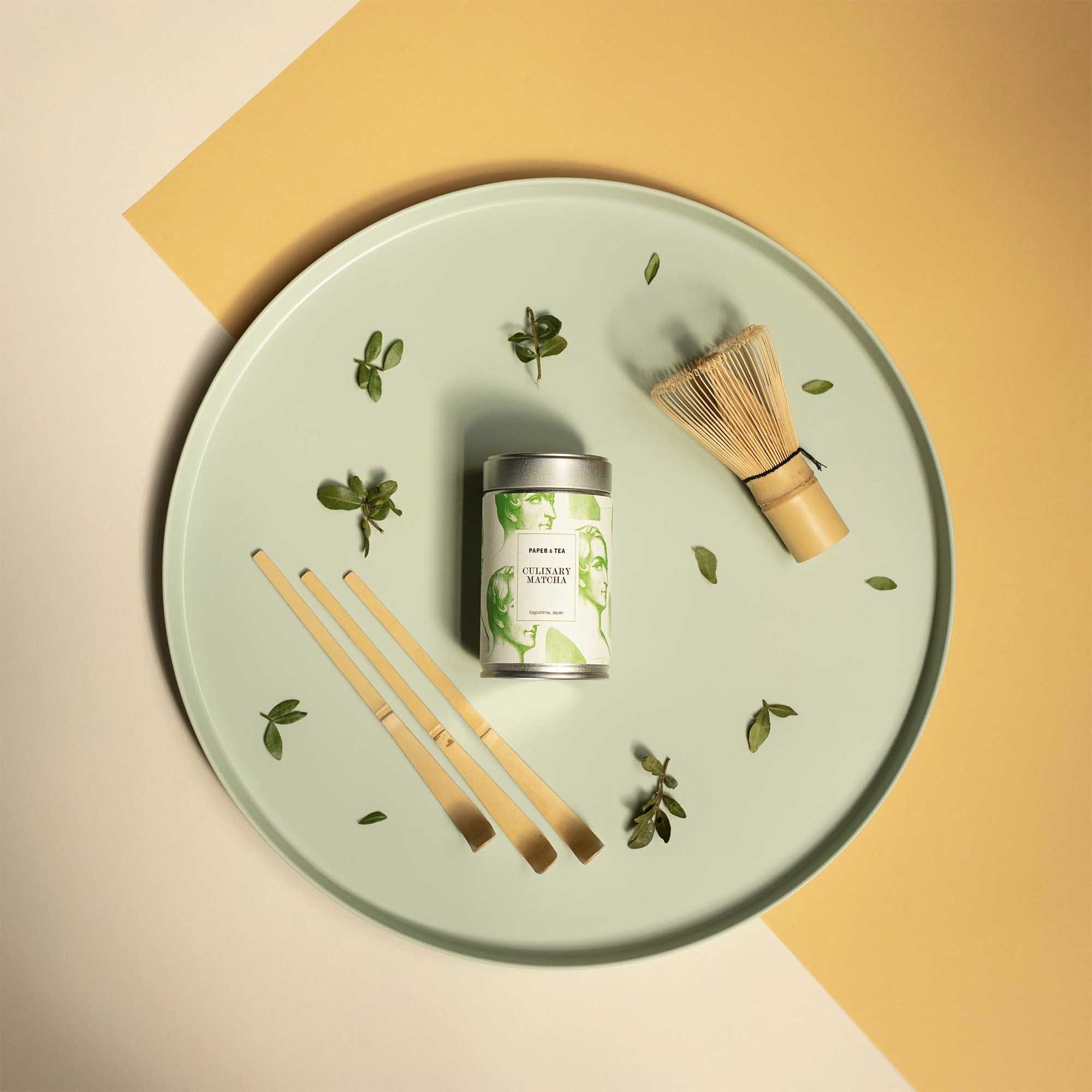 Rundes grünes Tablett mit PAPer & TEA Matcha-Dose, Bambus-Matcha-Besen, drei Messlöffeln und grünen Blättern.