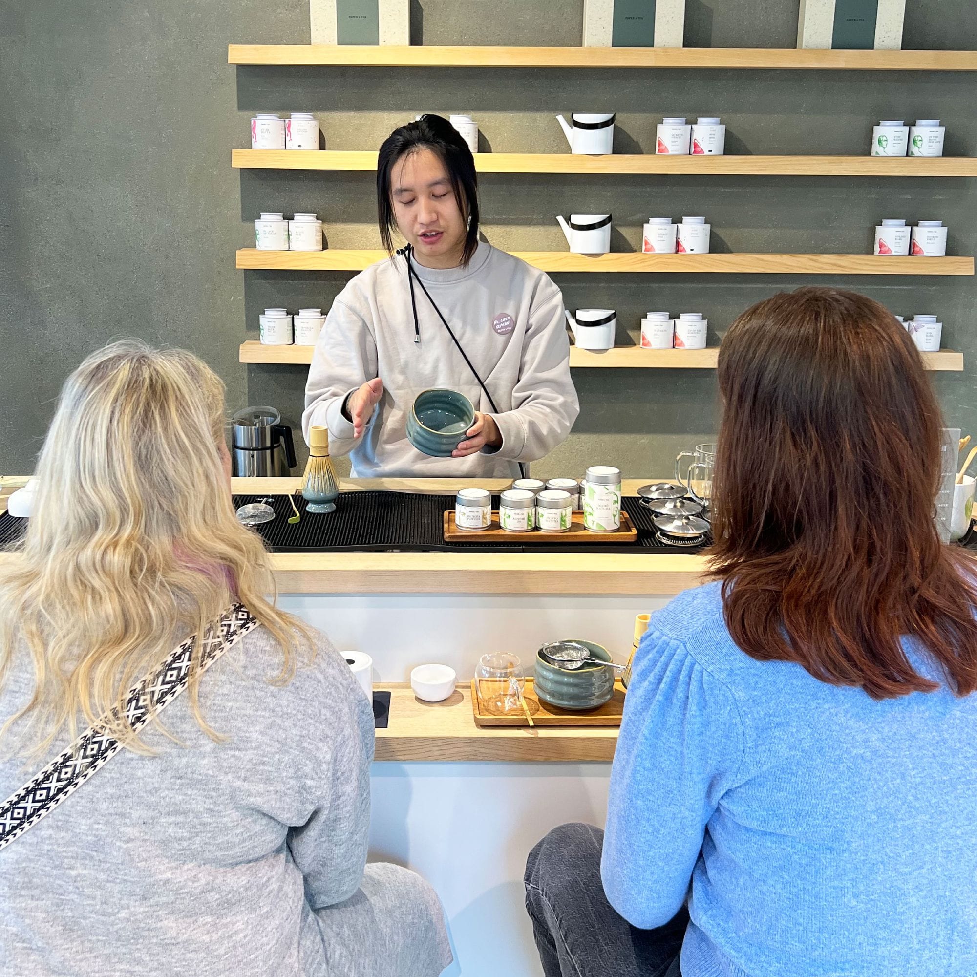 Mann in grauem Sweatshirt zeigt Teezubereitung mit PAPER + TEA Utensilien hinter Holztresen.