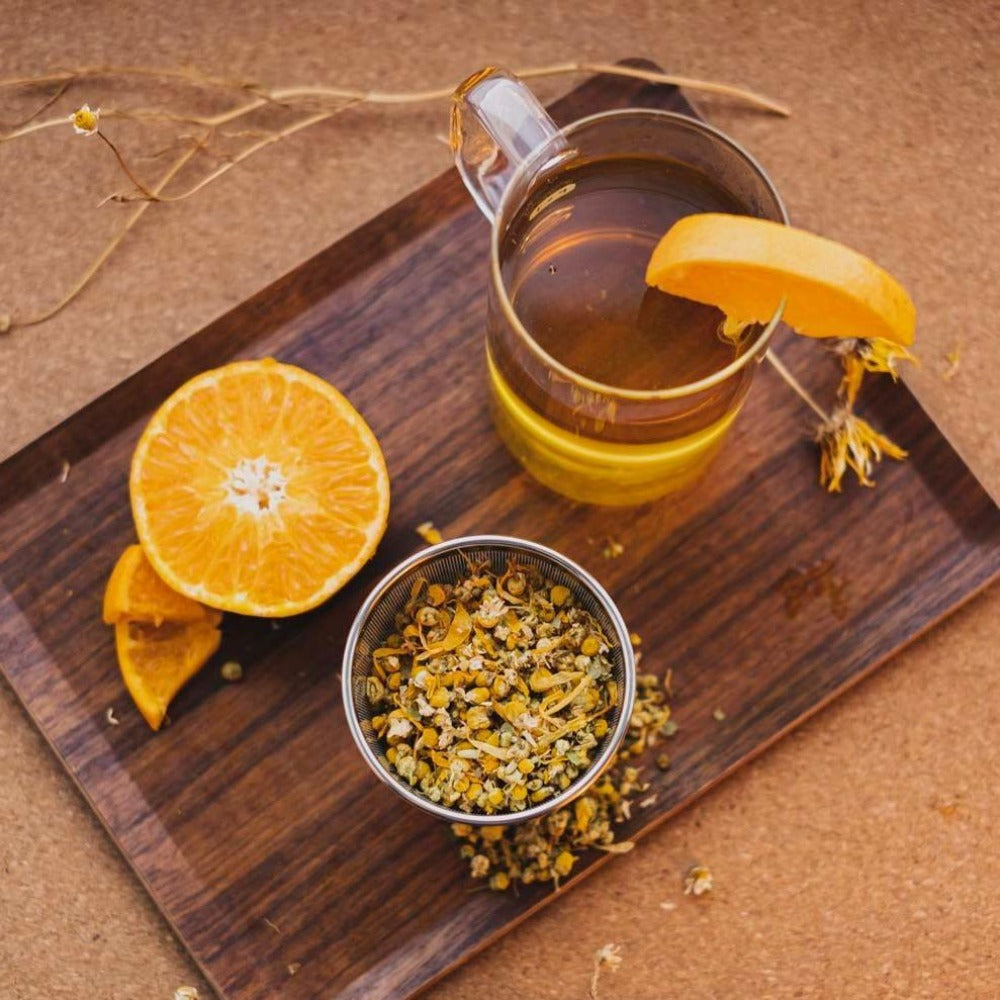 Glaskaraffe mit Tee, Orangenscheibe auf Holzbrett, halbe Orange, Kamillenblüten im Metallbehälter