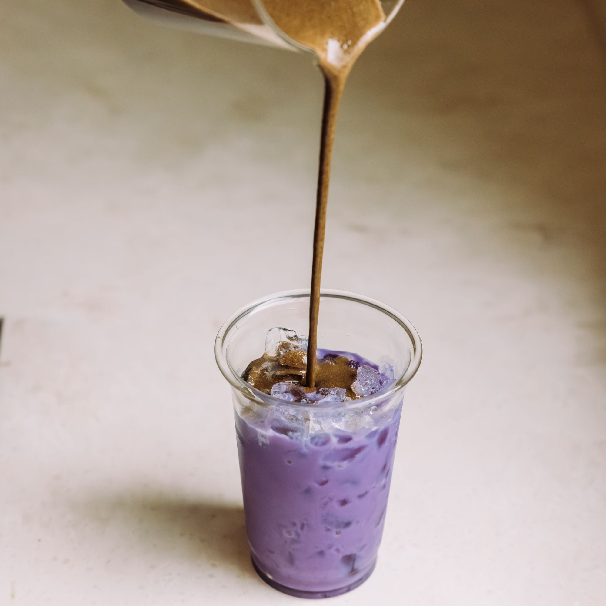 Klare Plastikbecher mit Eis und violettem Getränk, in das braune Flüssigkeit gegossen wird, auf beigem Hintergrund.