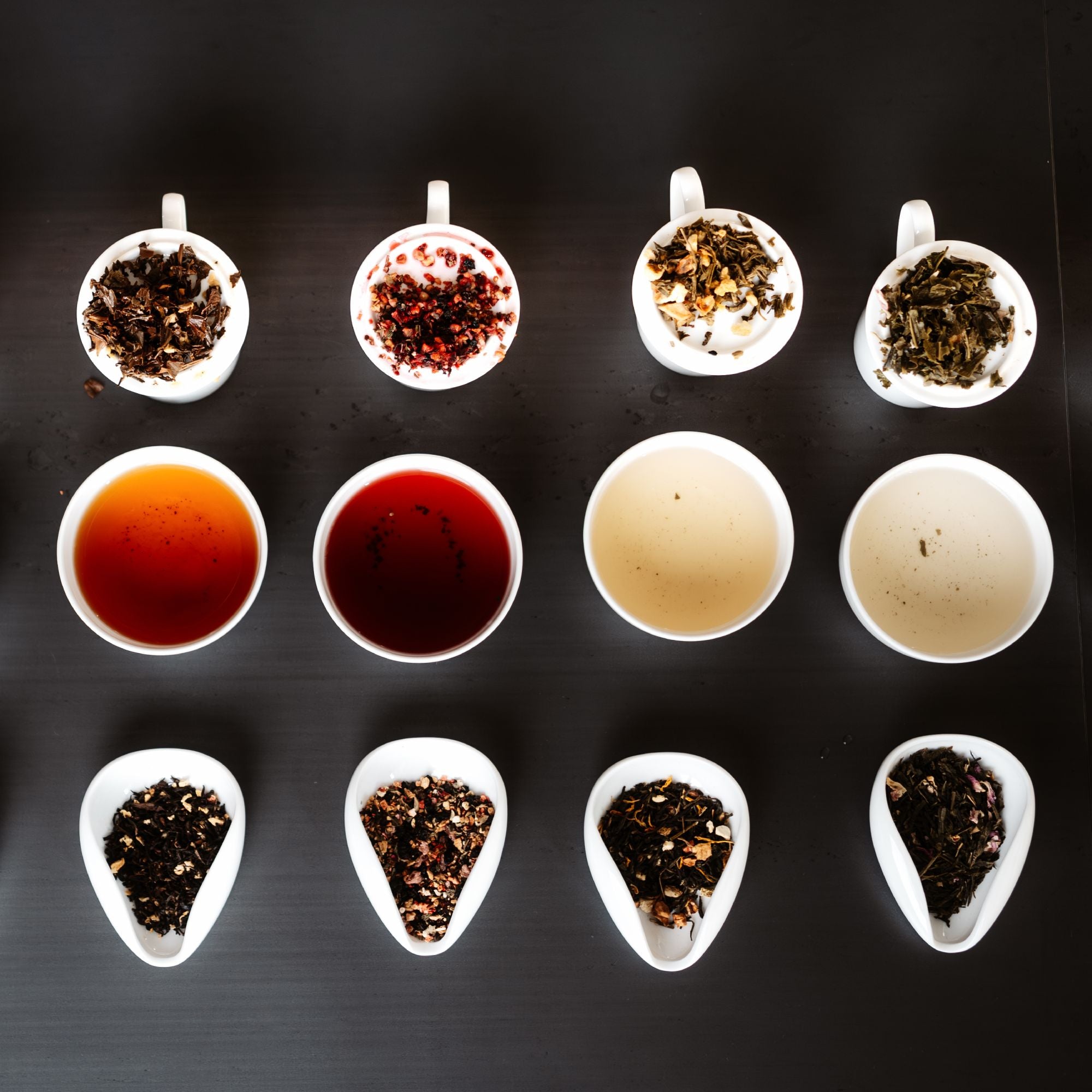 Vier weiße Tassen mit verschiedenen losen Teeblättern und Teefarben auf Holzuntergrund.