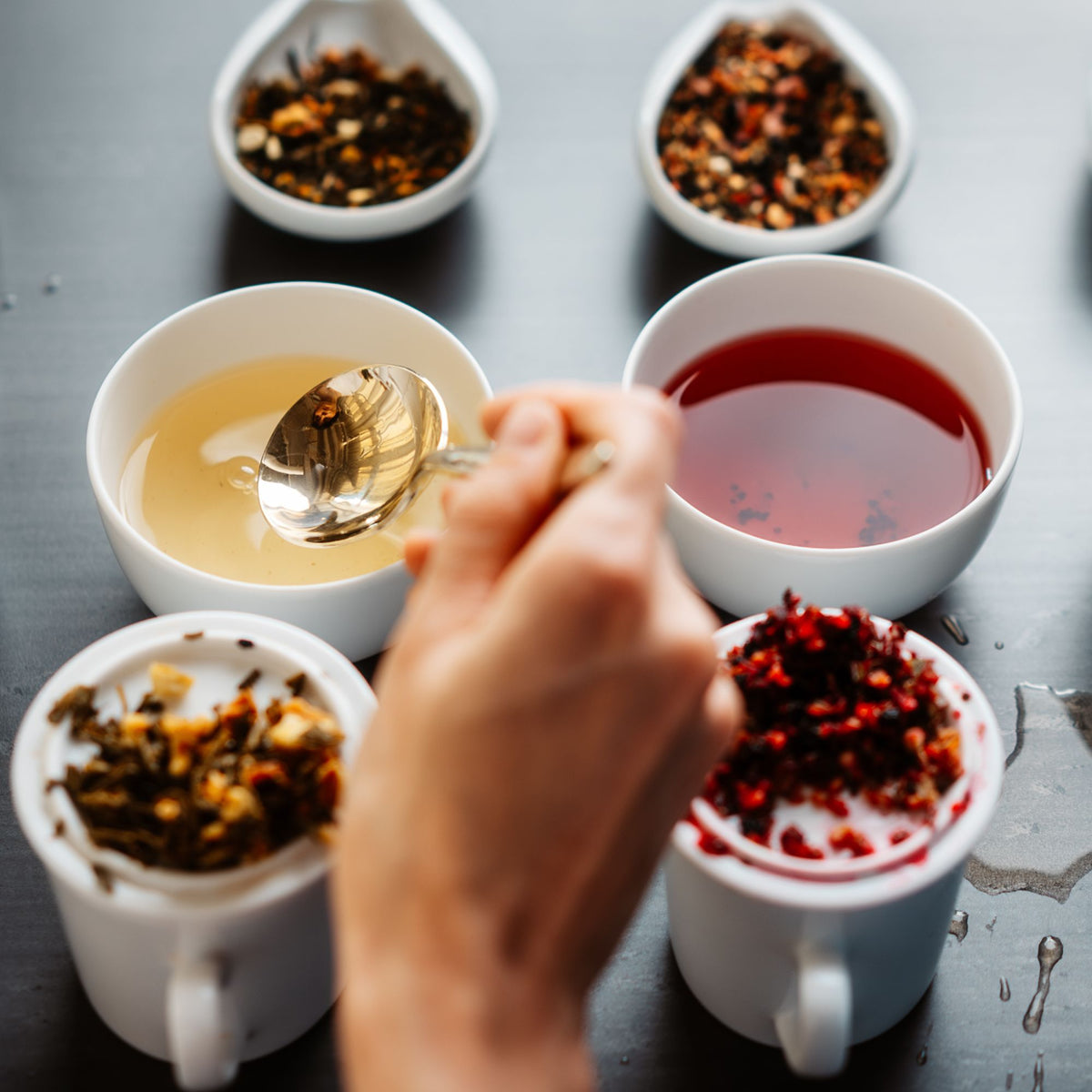 Hand hält silbernen Löffel über weißer Schale mit hellem Tee; daneben roter Tee und Becher mit Teeblättern.
