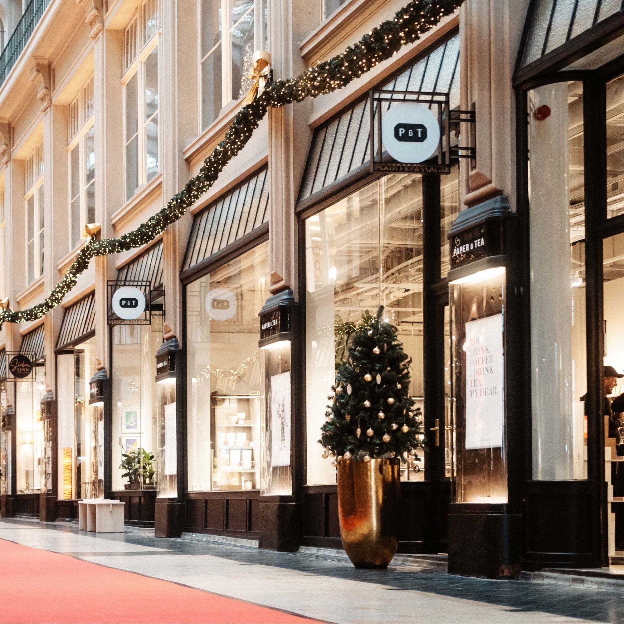 Weihnachtlich dekorierter PAPER & TEA Laden mit beleuchtetem Schaufenster und Tannenbaum in goldenem Topf