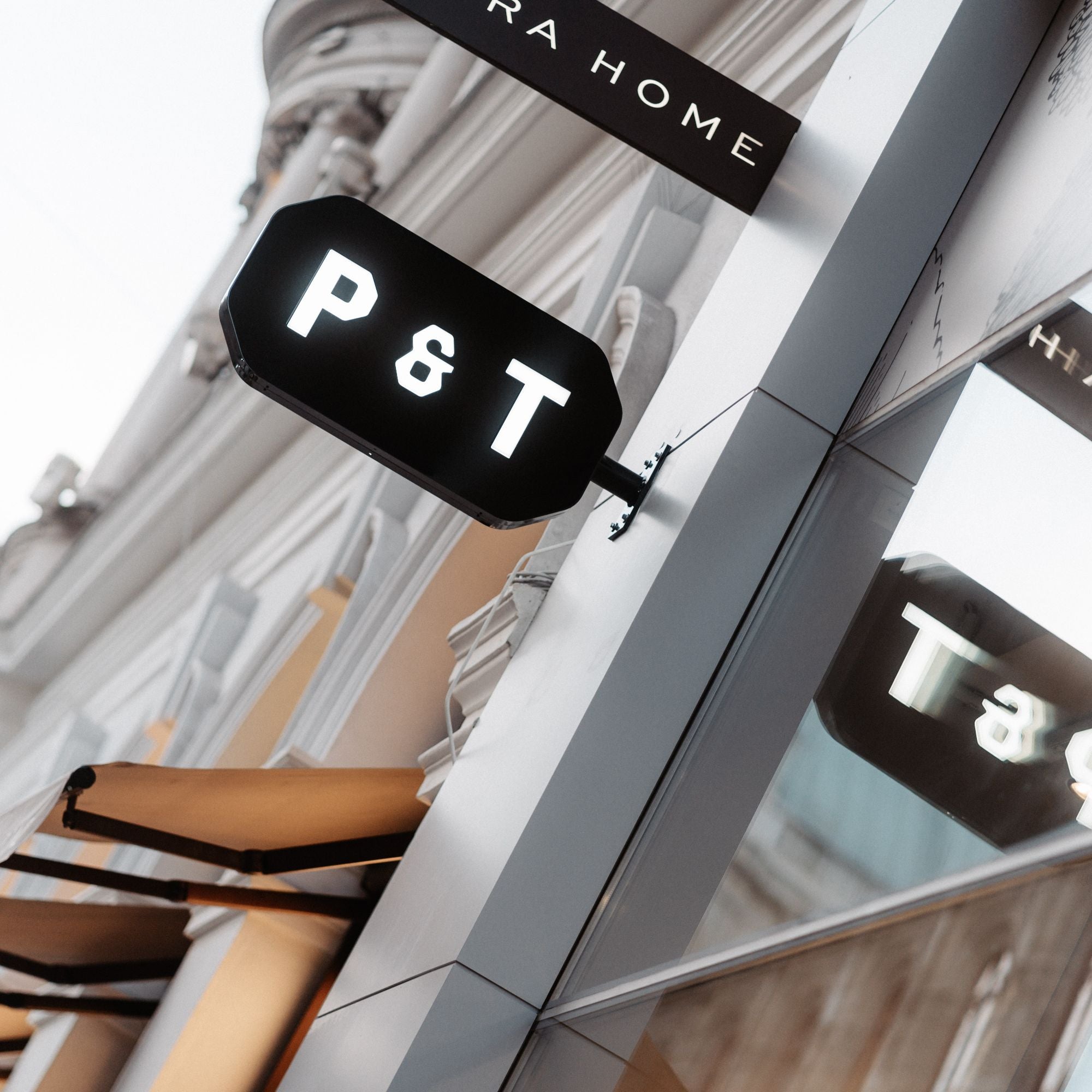 Schwarzes beleuchtetes P & T-Schild an moderner Gebäudeoberfläche mit braunem RA HOME-Banner darüber.