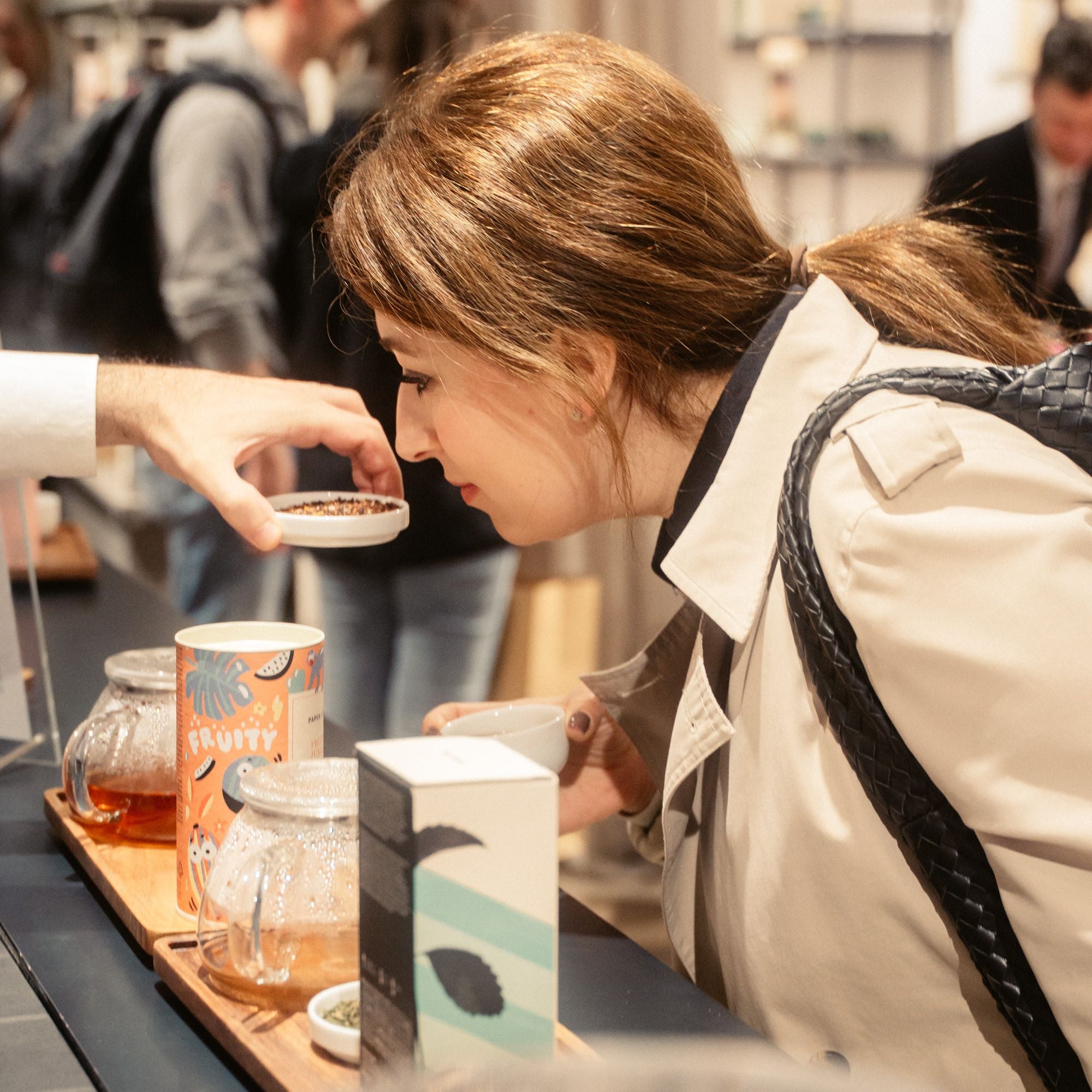 Frau im beigen Trenchcoat riecht an einem weißen Behälter mit Tee, bunte FRUITY-Teedose auf dem Tresen.
