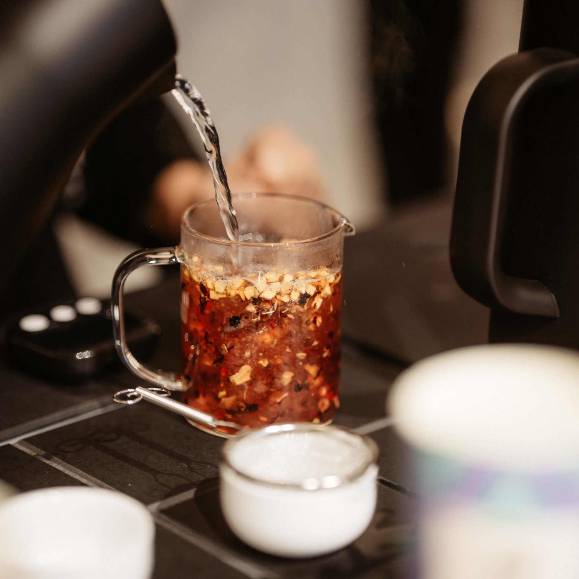 Heißes Wasser aus schwarzem Kessel wird in Glas mit dunklem Blüten- und Kräutertee auf Fliesenuntergrund gegossen.