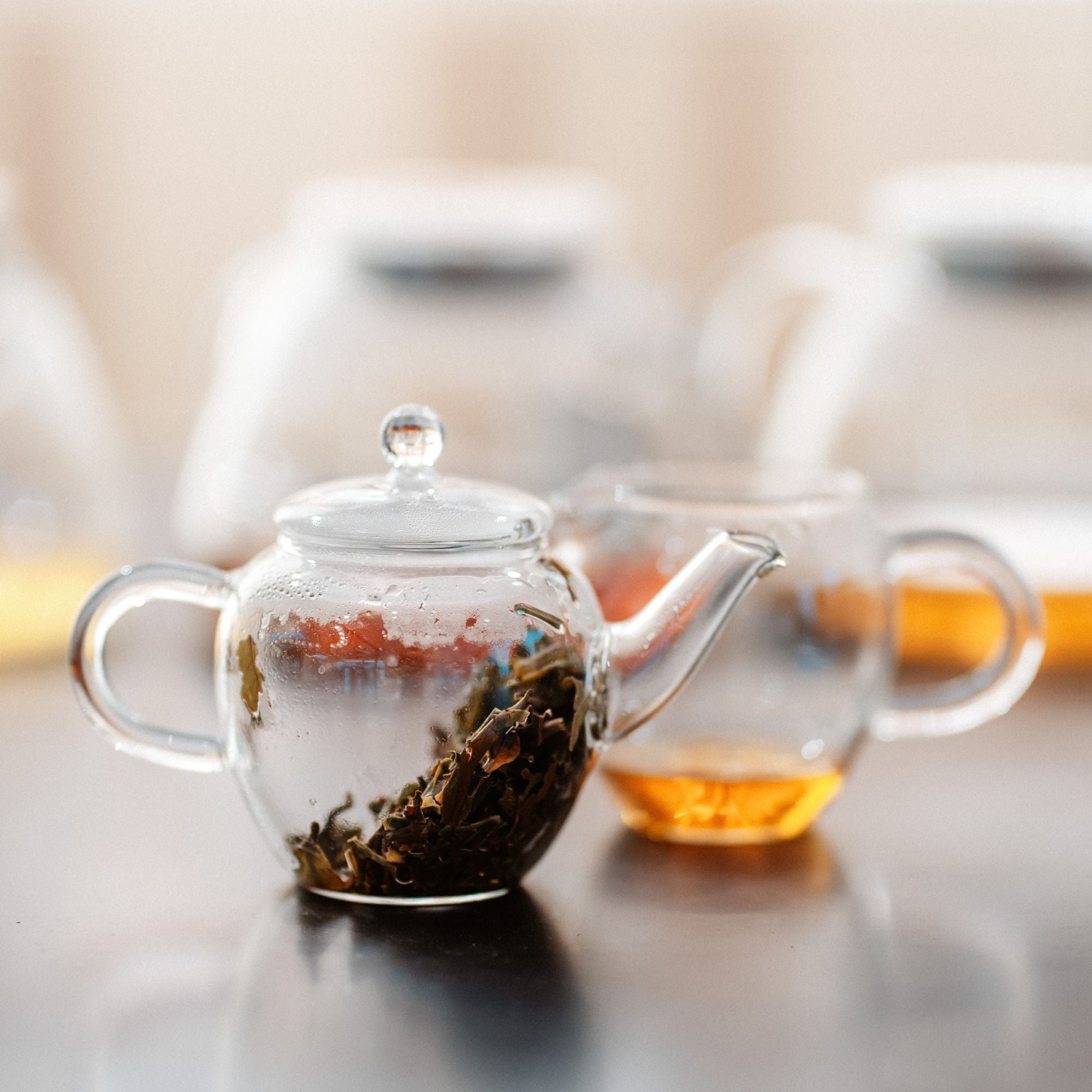 Klare Glas-Teekanne mit losen Teeblättern und Glastasse mit bernsteinfarbenem Tee auf dunklem Untergrund