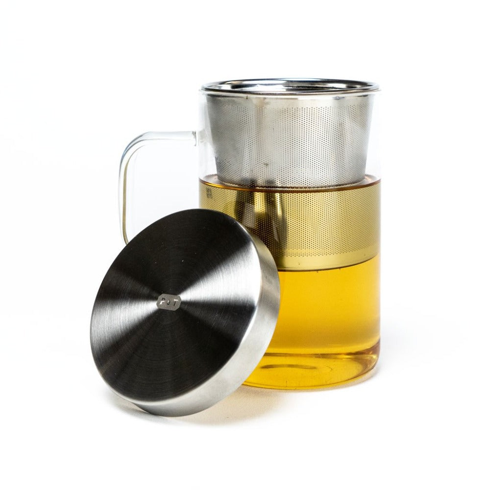 Transparenter Glasbecher mit goldgelbem Tee und feinem Metallsieb, davor ein Metalldeckel mit 'POT' Gravur.