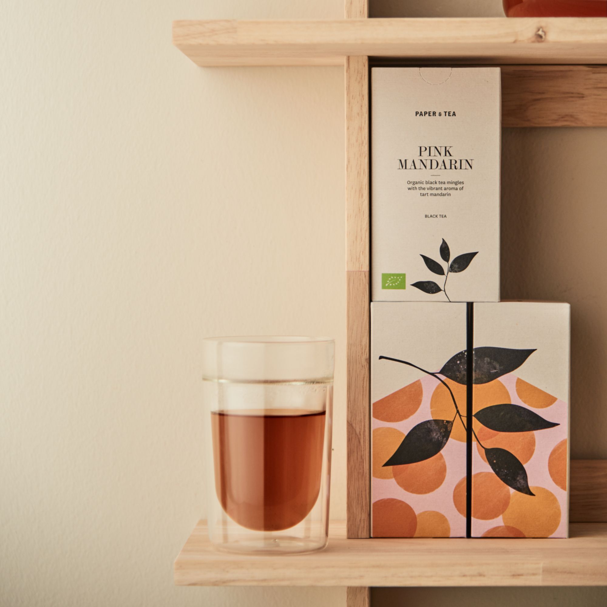 Paper & Tea Pink Mandarin Schwarztee Verpackung neben doppelwandigem Glas mit Tee auf Holzregal.
