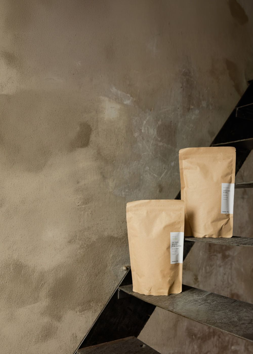 Zwei braune Papiertüten mit weißen 'IN THE MOOD FOR LOVE'-Etiketten auf Metalltreppe vor beiger Wand.