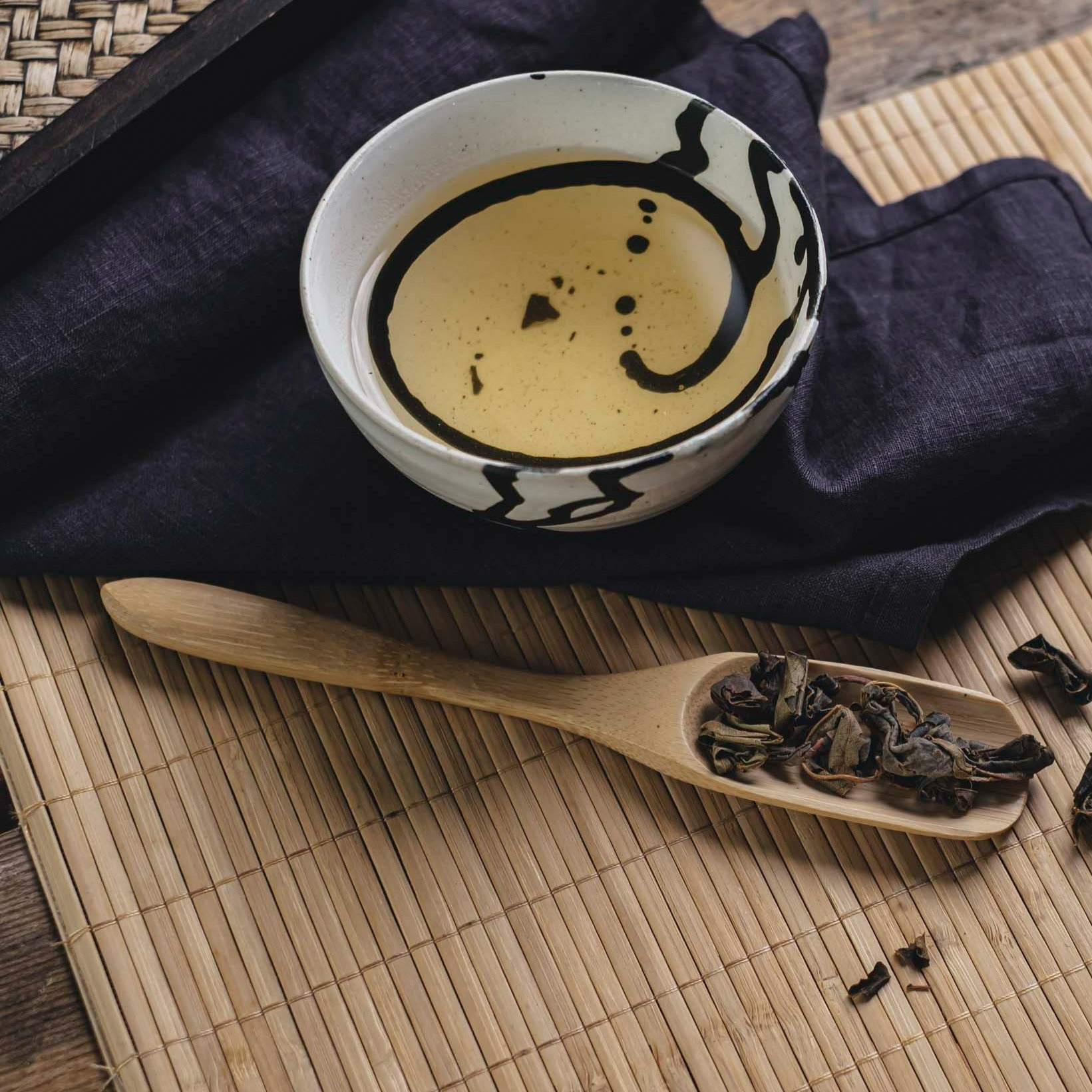 Weißer Keramikbecher mit schwarzem Muster, gefüllt mit hellem Tee, neben Holzlöffel mit Teeblättern auf Bambusmatte.