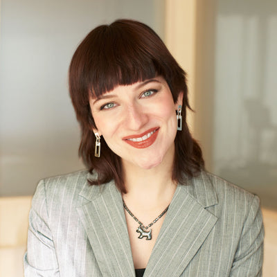 Junge Frau mit braunen Haaren, Ohrringen und Kette, im grauen Nadelstreifen-Blazer.