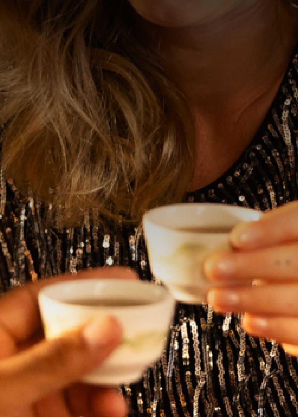 Zwei Hände halten kleine weiße Keramikbecher mit dunklem Tee vor Person in schwarzem Paillettentop.
