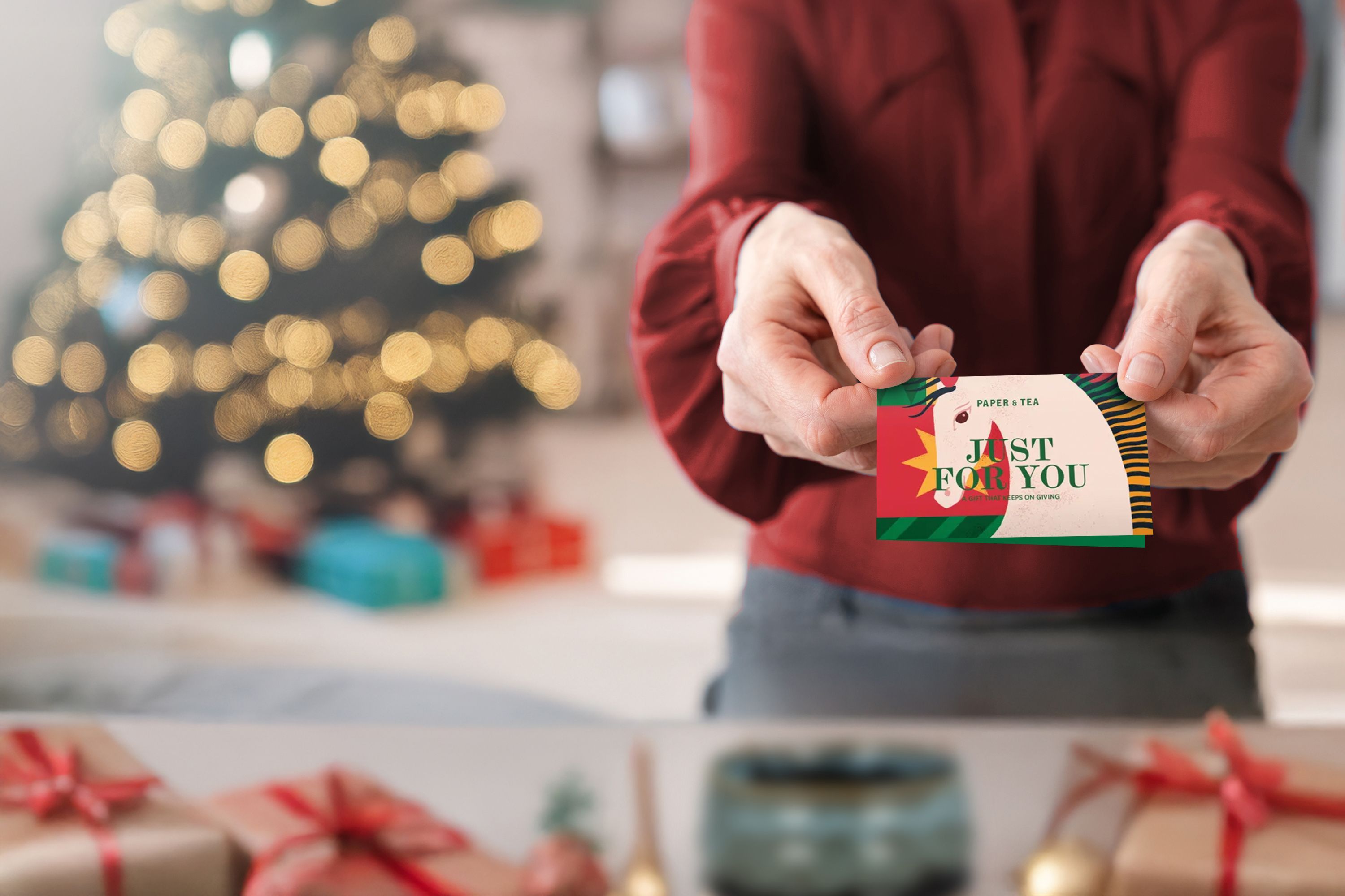 Person hält bunte PAPER & TEA Geschenkkarte vor Weihnachtsbaum mit Lichtern und Geschenken mit roten Schleifen.