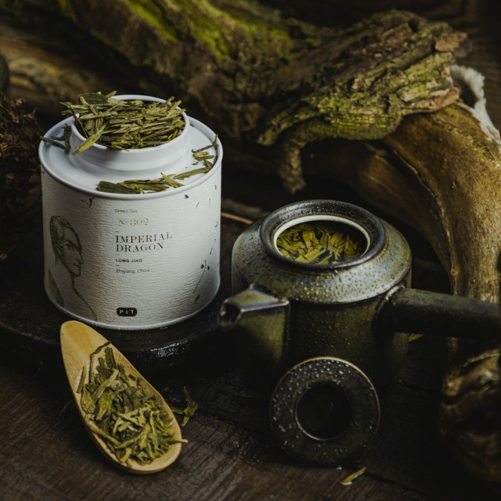 Weiße Dose Imperial Dragon Long Jing Grüntee, grüner Teekrug mit Blättern und Holzlöffel im rustikalen Ambiente.