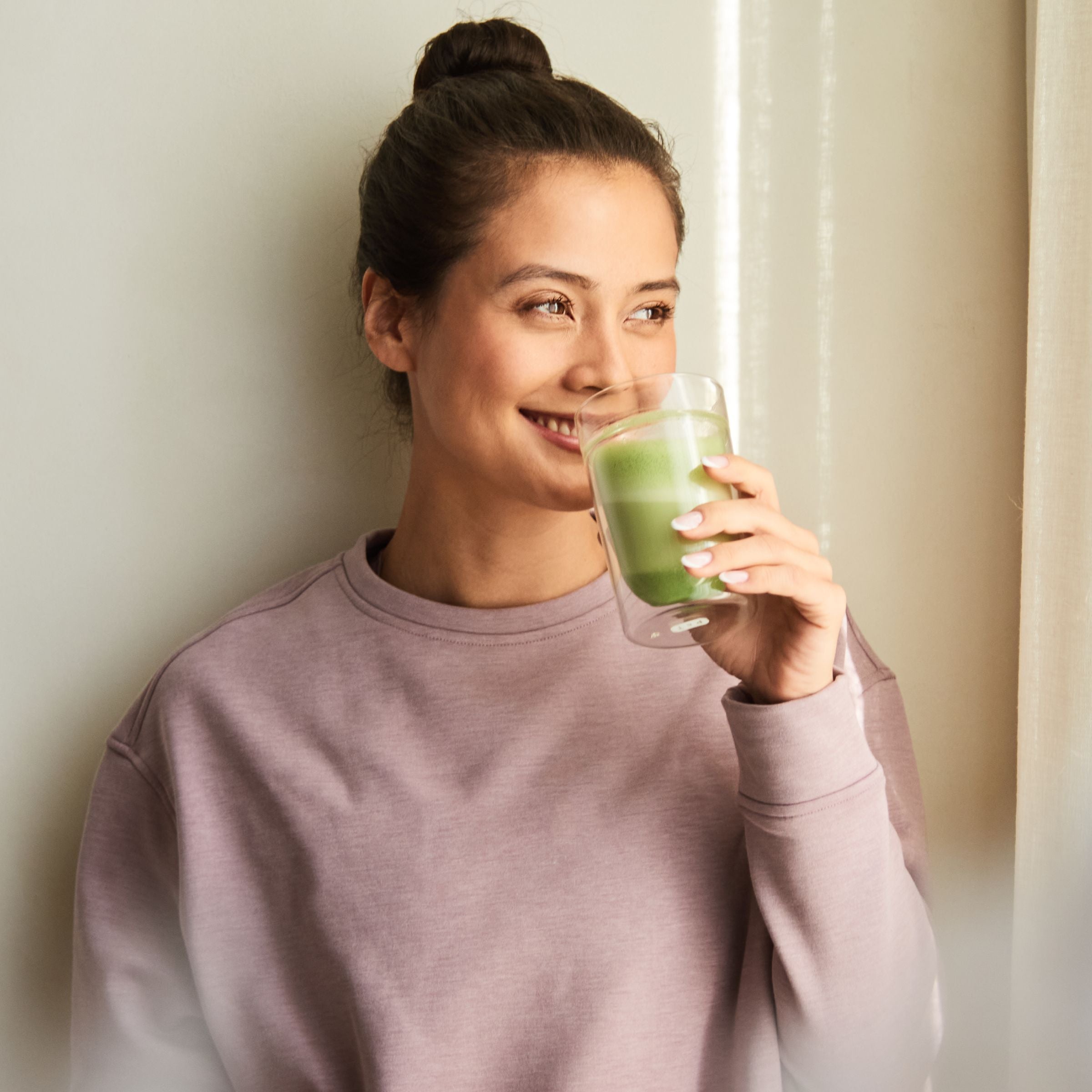 Frau mit braunem Dutt und lila Sweatshirt trinkt grünes Getränk aus klarem Glas in ruhiger Innenatmosphäre.