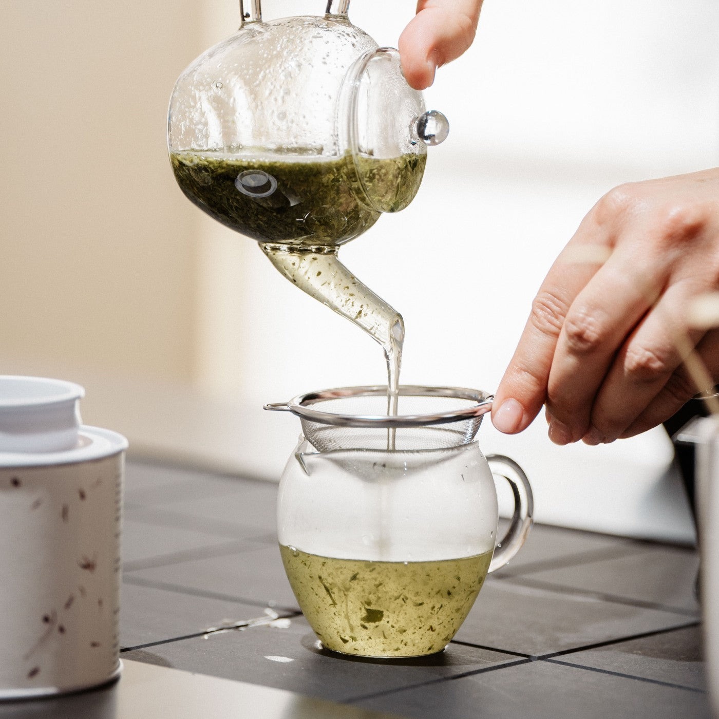 Glasteekanne gießt grünen Tee durch Metallsieb in Glasbecher auf dunklem Tisch neben Keramikbehälter.