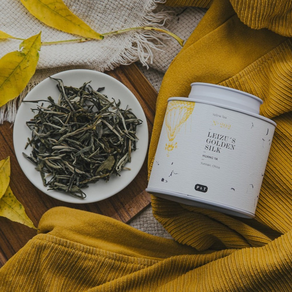 Weiße Platte mit getrockneten gelben Teeblättern und Teebehälter auf gelbem Stoff, Golden Silk Huang Ya Tee.
