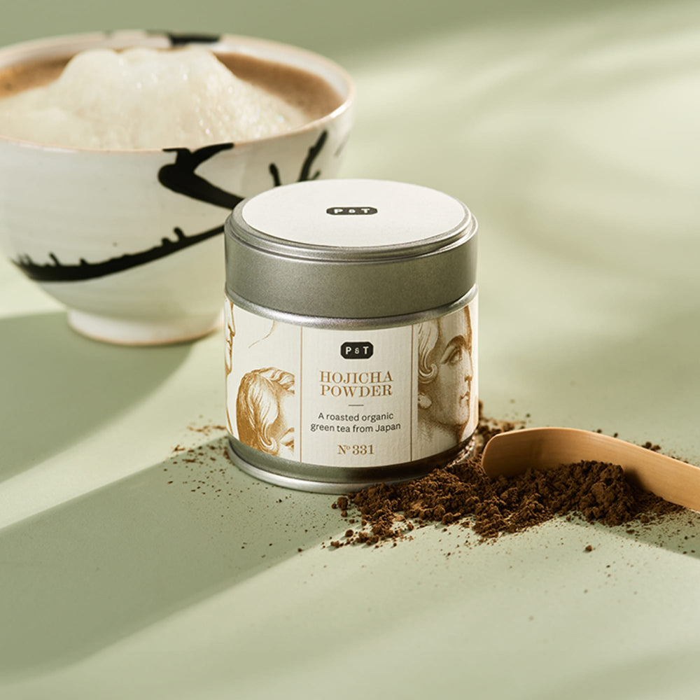 Zylinderförmige Metalldose mit Etikett für Hojicha Grünteepulver, Holzlöffel mit Pulver und Keramikschale mit Getränk.