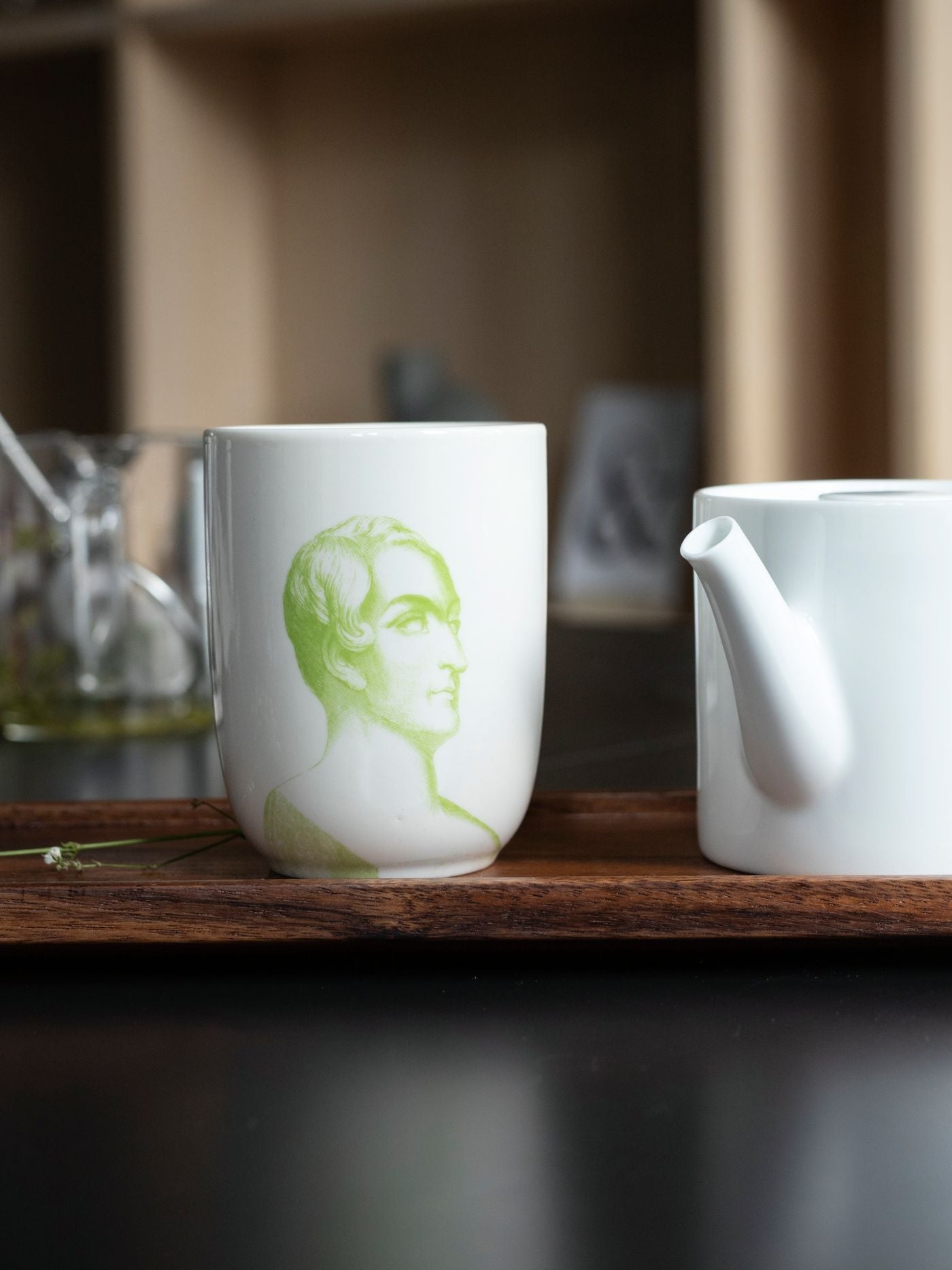 Weißer Keramikbecher mit grünem Männerprofil auf einem Holztablett neben weißem Teekännchen.