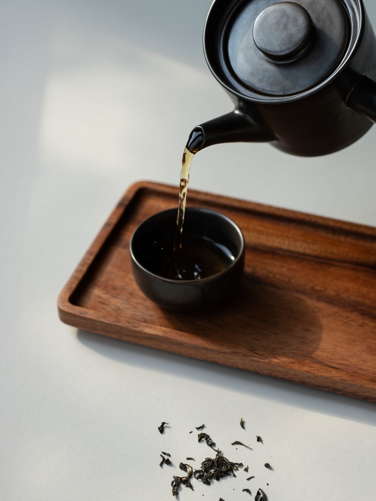 Schwarze Teekanne gießt Tee in kleine schwarze Tasse auf Holztablett mit losen Teeblättern auf weißer Oberfläche.