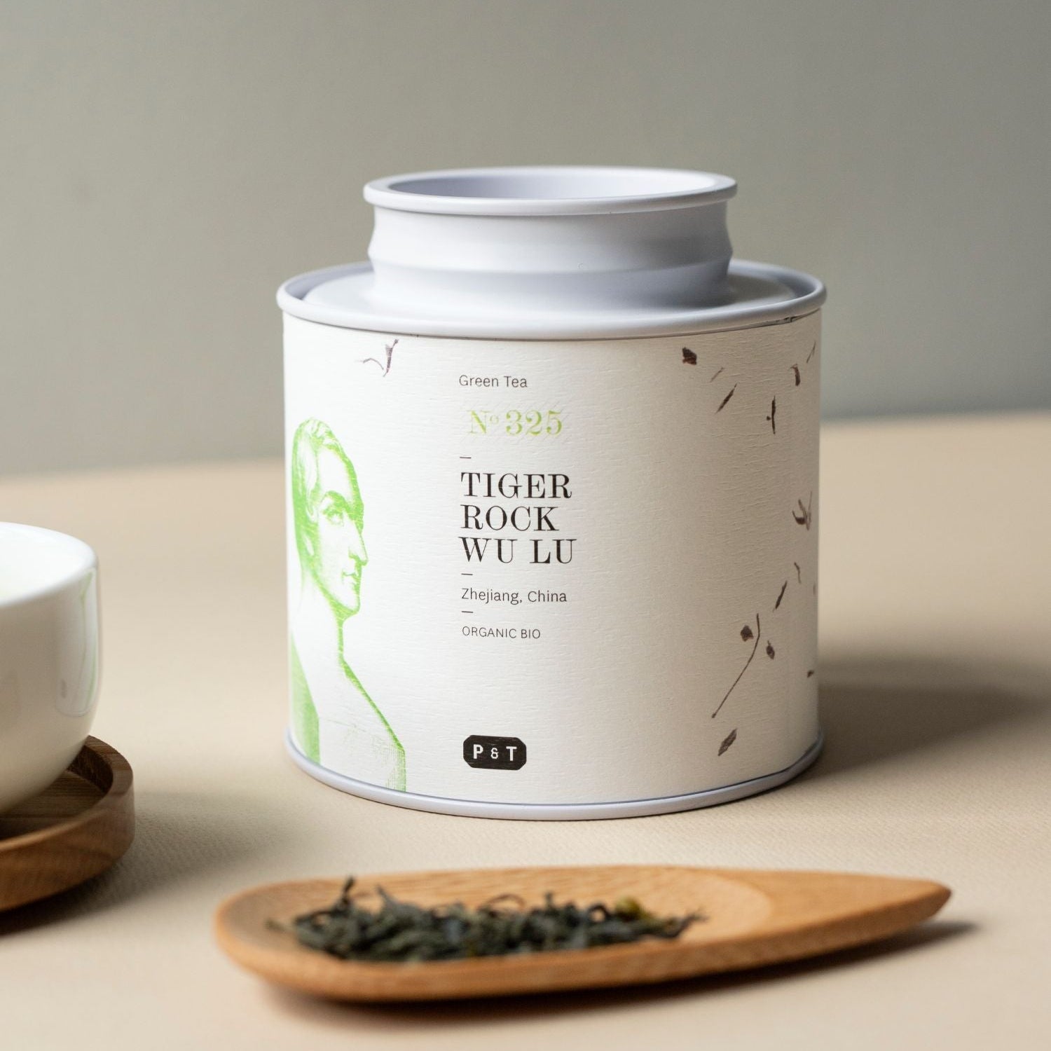 Weißer Teebehälter mit grünem Label ‚BIO-Tiger Rock Wu Lu‘, lose Blätter auf Holzlöffel, neben weißer Tasse.