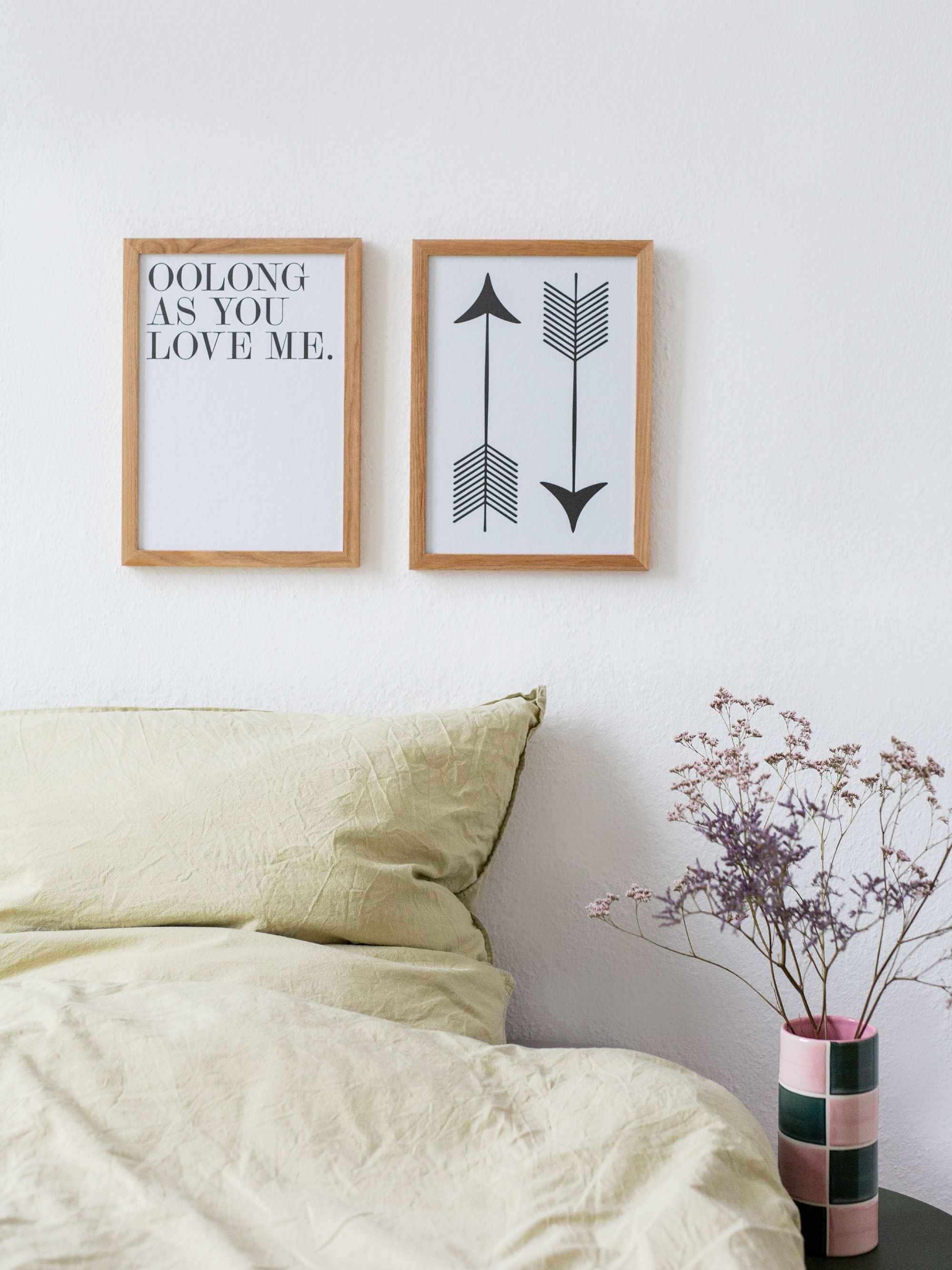Minimalistisches Schlafzimmer mit zwei Holzrahmenbildern, Schriftzug 'OOLONG AS YOU LOVE ME', cremefarbener Bettwäsche.