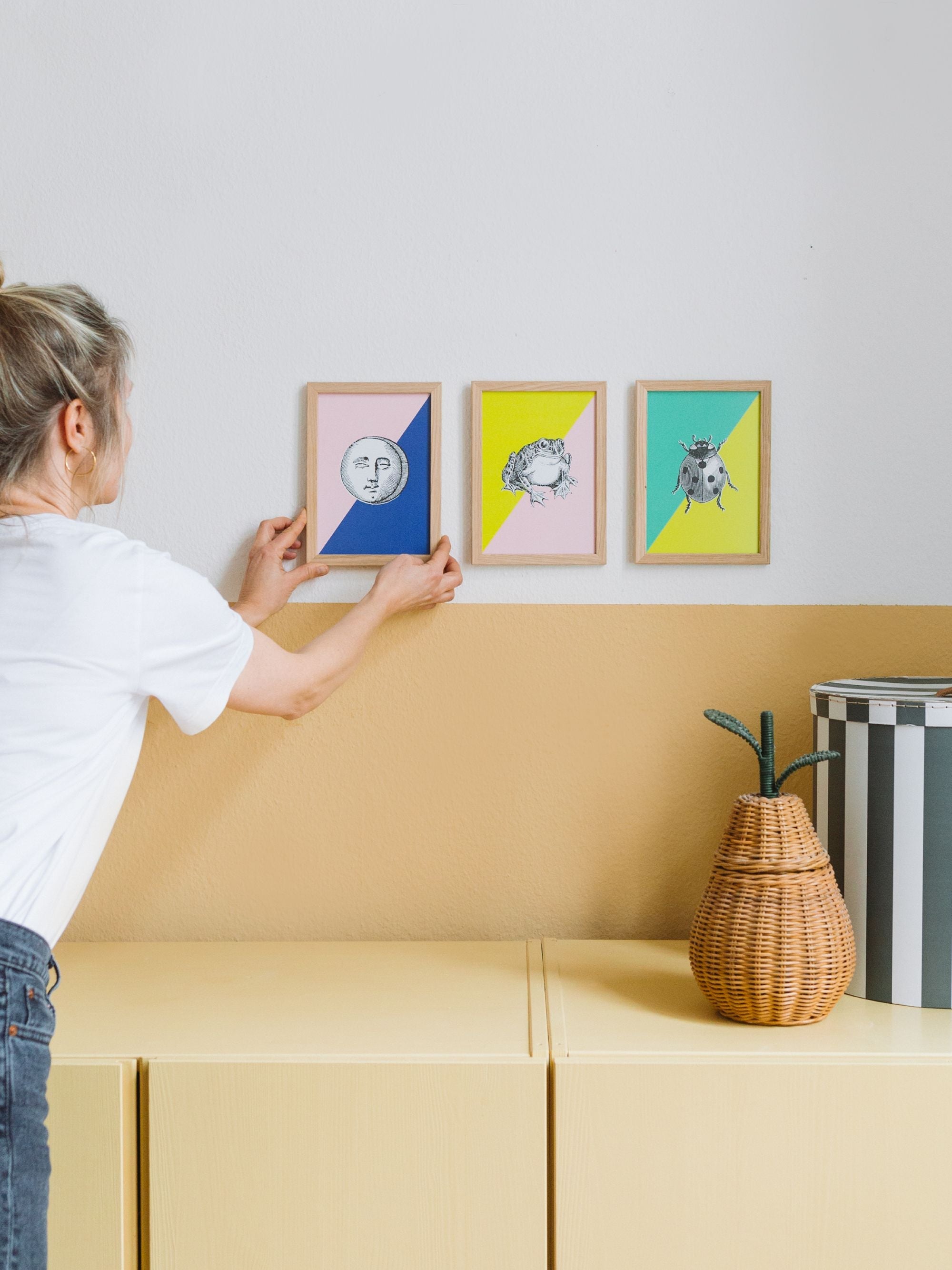 Frau hängt gerahmtes Kunstbild mit Mondgesicht und Frosch über gelben Schrank mit Korb auf.