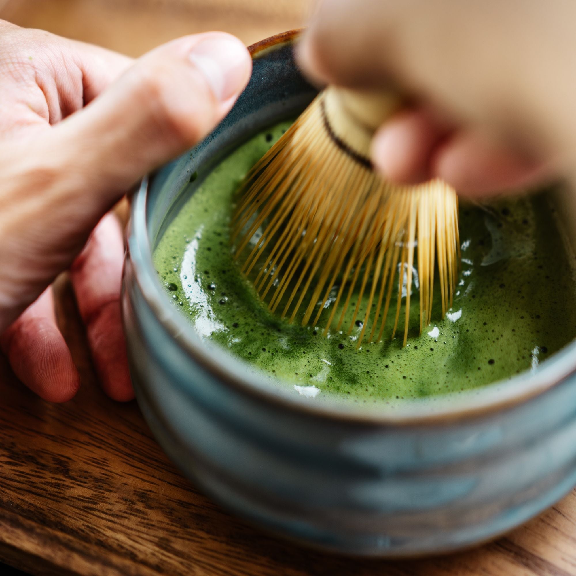 Hände schäumen leuchtend grünen Matcha-Tee mit Bambusbesen in einer blauen Keramikschale auf Holzuntergrund.
