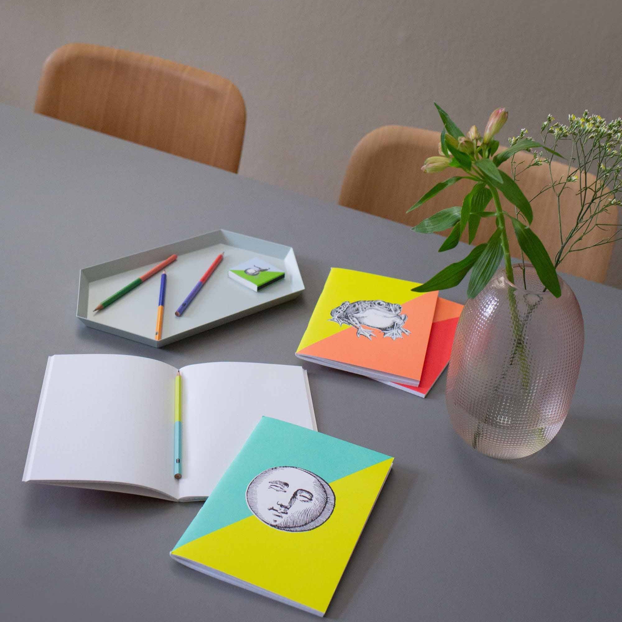 Grauer Tisch mit bunten Notizbüchern, offenem Notizbuch, Farbstiften im Tablett, Vase mit Blumen und Holzhockern.