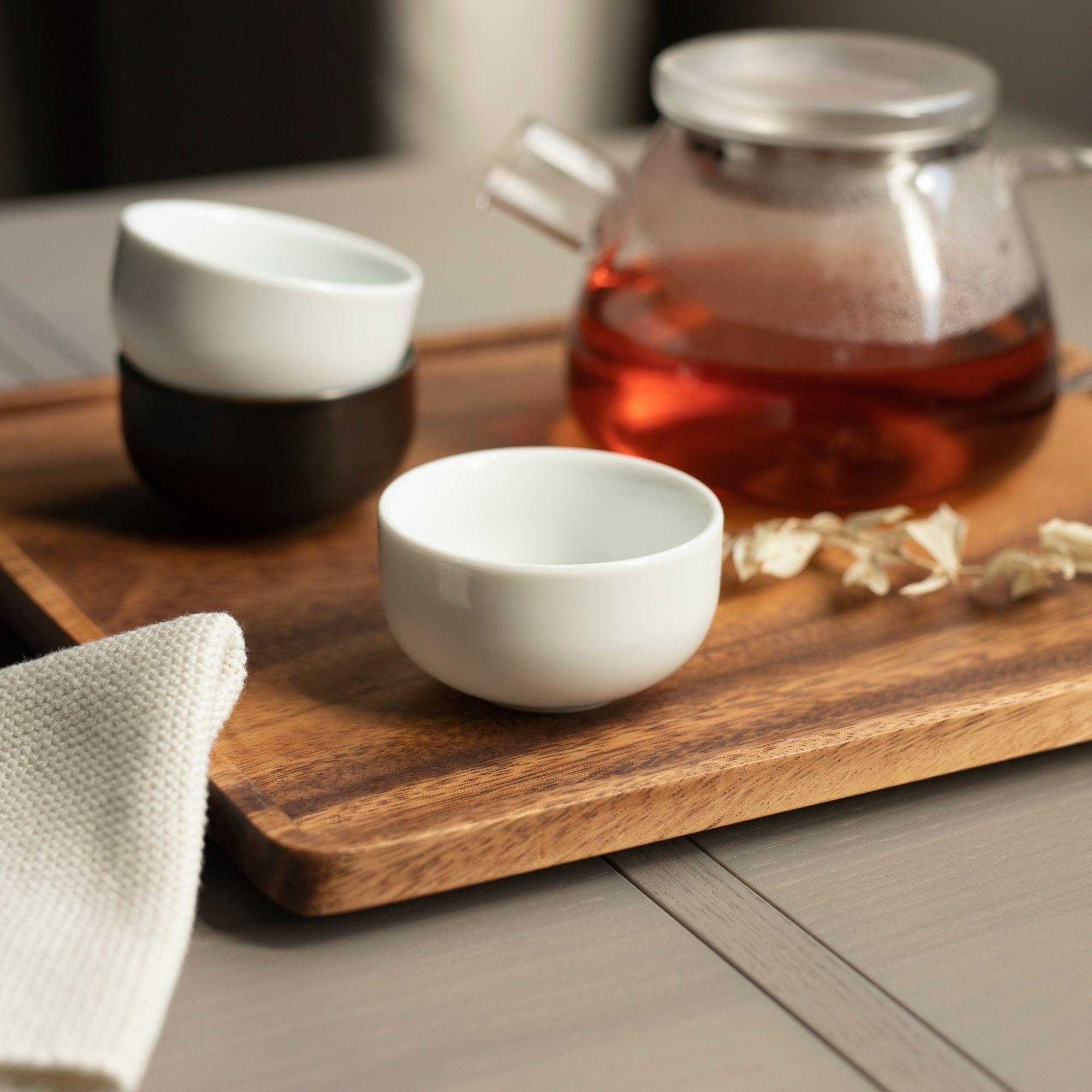 Glaskanne mit herbstlichem Tee auf Holztablett neben weißen und schwarzen Keramiktassen, dekoriert mit getrockneten Blüten.