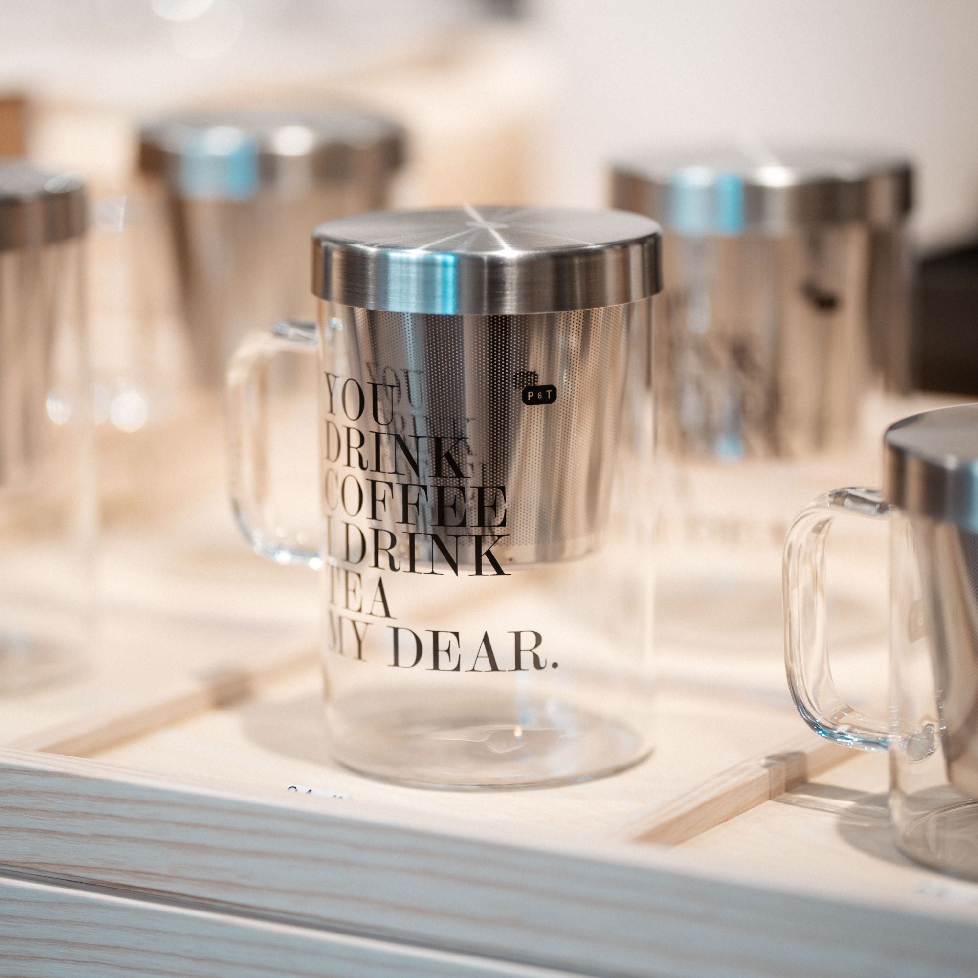 Transparenter Glasbecher mit Metallfilter und schwarzem Schriftzug auf einem hölzernen Tablett.