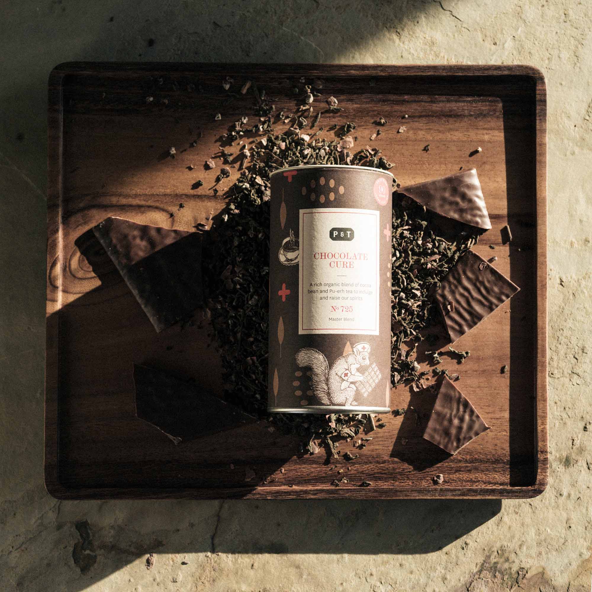 Zylindrische Dose 'P & T CHOCOLATE CURE', umgeben von Pu-erh-Teeblättern und dunkler Schokolade auf Holztablett.