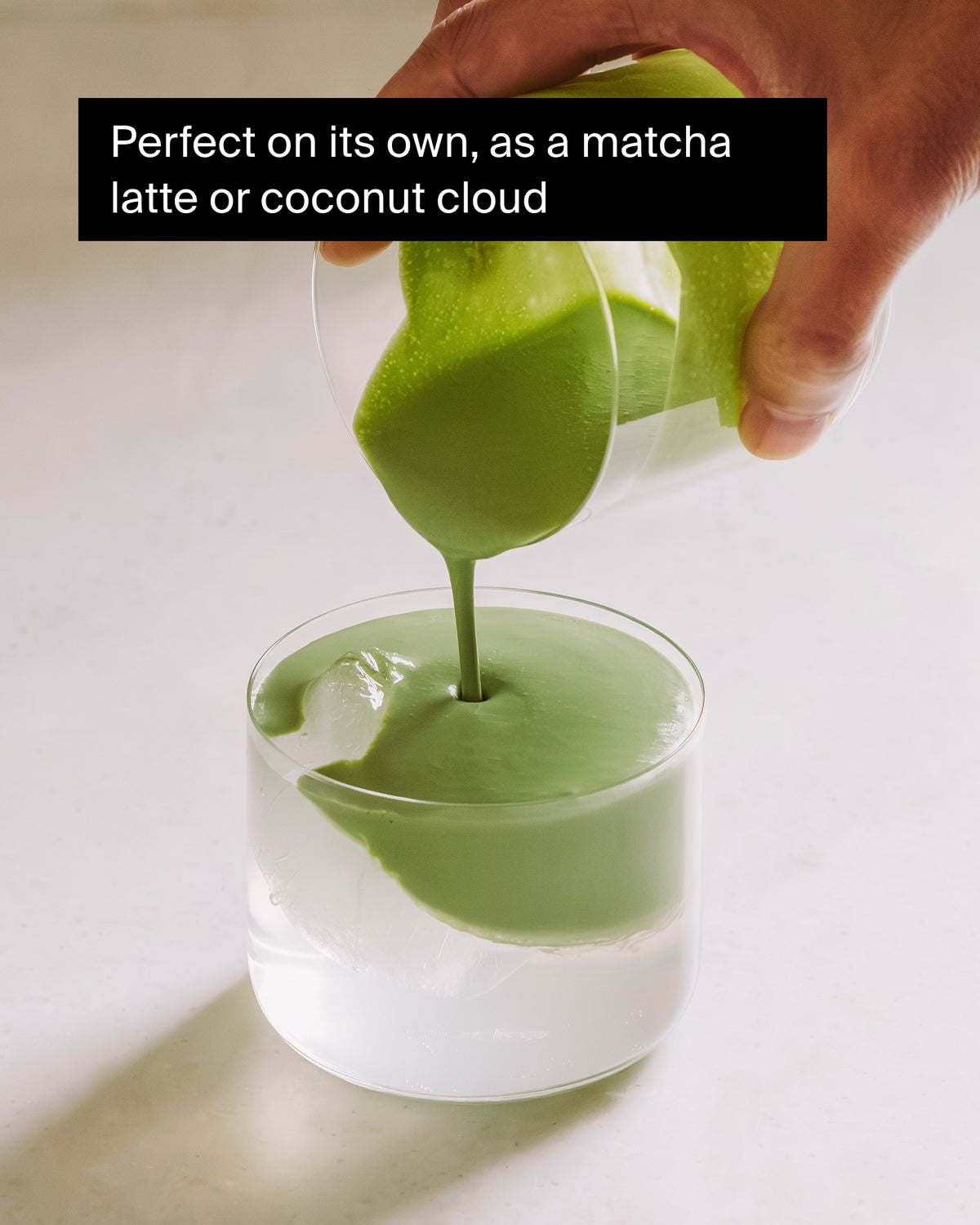 Hand gießt cremigen Matcha-Latte in ein Glas mit Eiswürfeln auf hellem, neutralem Hintergrund.