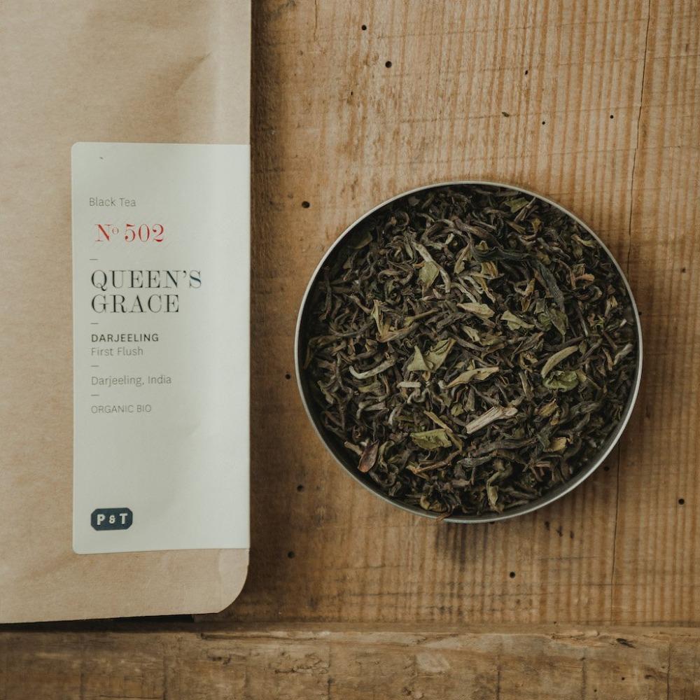 Beige Papiertüte mit Bio-Darjeeling Schwarztee 'Queen's Grace' neben einem Metallgefäß mit losen Teeblättern.